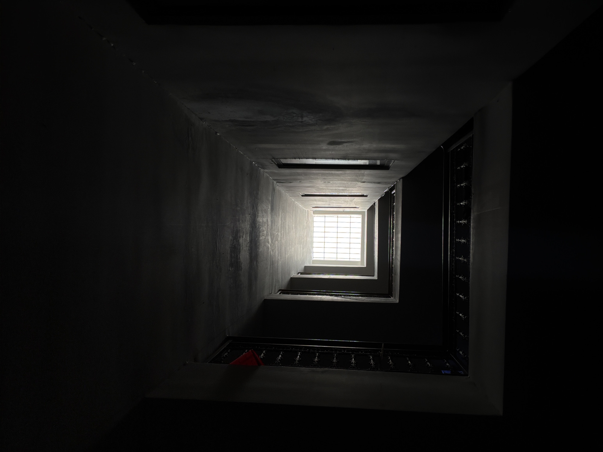 The image depicts a view looking upwards through a stairwell. The walls are concrete, with varying degrees of light exposure, appearing dark in the foreground and progressively brighter towards the top. A square window with a grid pattern is visible at the top of the stairwell, allowing light to penetrate. Staircases are discernible, with railings featuring decorative elements. A red object is seen on one of the lower landings. The overall environment is dimly lit. No people are visible. There is no evidence to confirm that this picture was taken in Đà Nẵng, Vietnam.
