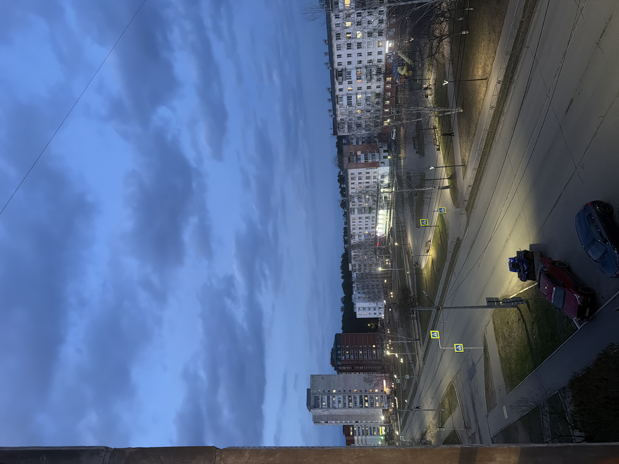 The image captures an urban landscape at dusk or early night, likely from an elevated vantage point such as a balcony or window, overlooking a street in Chaikovsky, Russia. The sky above is a deep, cloudy blue, with the last hints of daylight fading.
In the foreground, a concrete ledge borders the bottom of the frame. Beyond it, a wide road, potentially with multiple lanes, stretches across the scene. It is flanked by sidewalks and grassy verges. Numerous streetlights emit a warm yellow light, illuminating the thoroughfare and casting long shadows. Several yellow square signs featuring a black pedestrian crossing symbol are visible, marking designated crossings. Overhead wires, possibly for power or public transport, crisscross the upper sky.
Along both sides of the street and receding into the distance are various multi-story apartment buildings. Their architectural styles vary, with some appearing older and lighter in color, while others are newer with darker facades. Many windows within these buildings are lit, indicating occupancy. Bare trees are scattered throughout the scene, suggesting a late autumn or early spring season.
A few vehicles are present on the side of the road, including a dark blue car and a red car. Next to the red car, a blue all-terrain vehicle (ATV) is visible on a trailer. A lone figure can be seen walking on a sidewalk in the mid-ground, near what appears to be a children's playground structure. The overall atmosphere is quiet and serene, characteristic of an urban residential area as night falls. No specific text is clearly legible within the image.