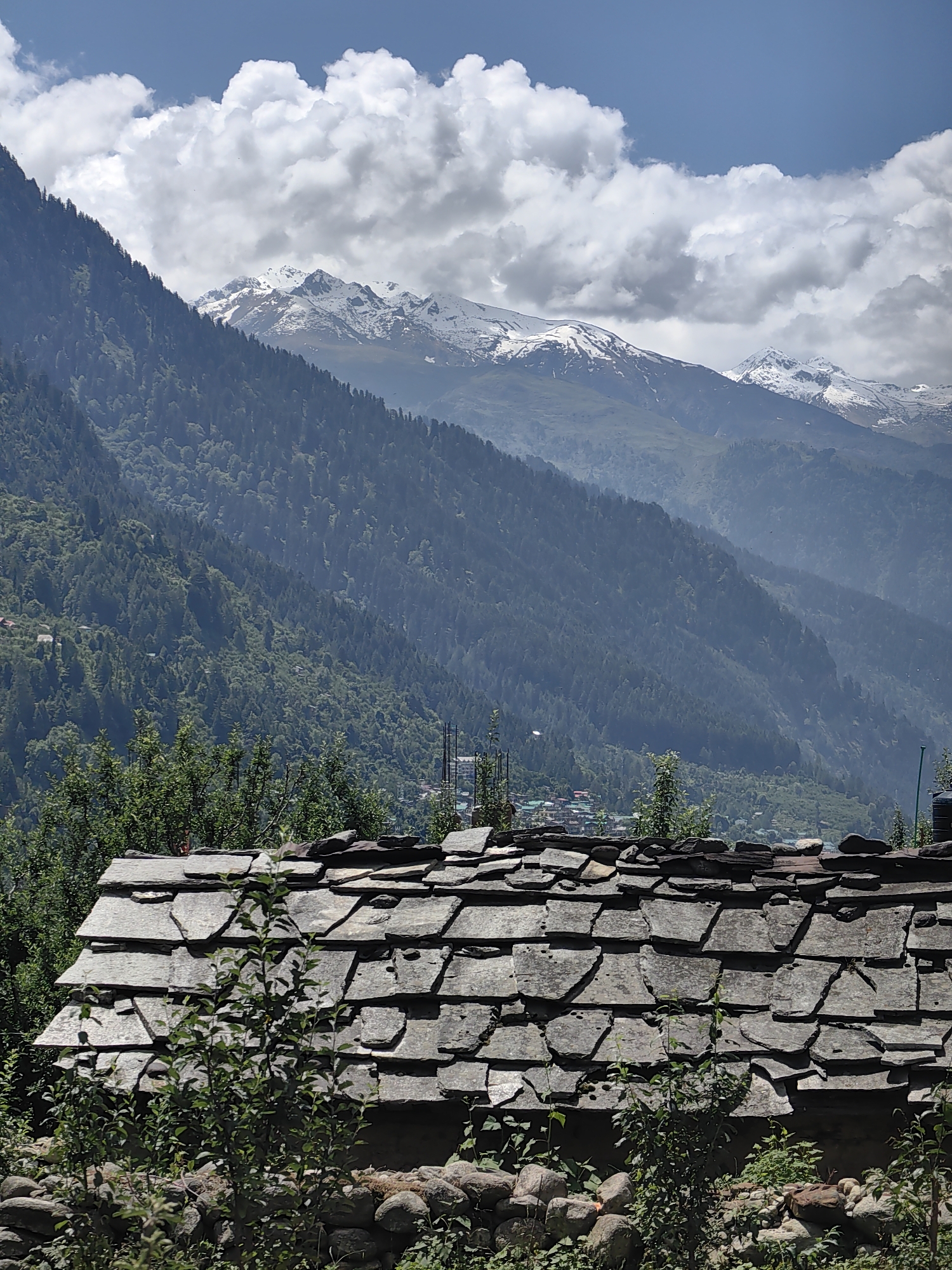 This image shows a close-up view of a traditional stone-tiled roof in the foreground, with lush green trees partially obscuring the view. In the mid-ground, a dense forest covers the slopes of a mountain, with a small village nestled amongst the trees. The background features majestic snow-capped mountains under a partly cloudy sky. The scene depicts a rural mountain landscape, likely in the Himalayas, consistent with the general geography of Barheta, India. No people are visible in the image.