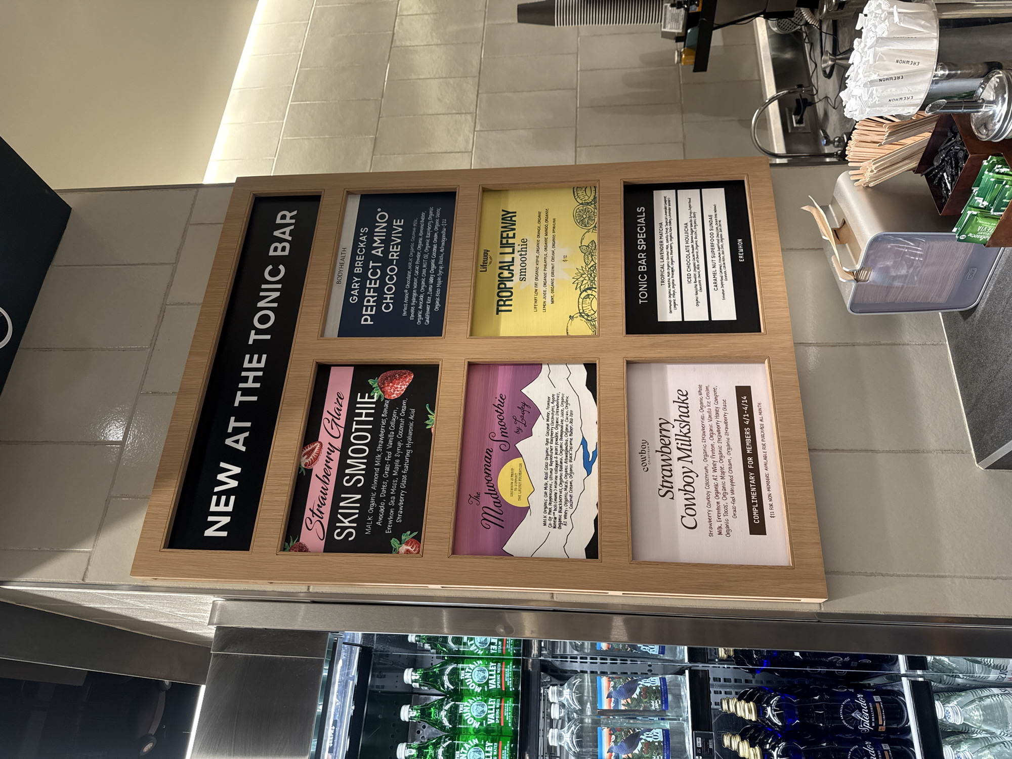 A framed menu board displaying smoothie and tonic bar specials is mounted above a refrigerated display case. The menu features six options: "Strawberry Glaze SKIN SMOOTHIE", "GARY BRECKA'S PERFECT AMINO CHOCO-REVIVE", "TROPICAL LIFEWAY smoothie", "TONIC BAR SPECIALS", "The Madwoman Smoothie by Laufey", and "Cowboy Strawberry Cowboy Milkshake". Each option is described with its ingredients. The refrigerated case below contains bottles of water. The background shows a tiled wall and glimpses of beverage preparation equipment. The location is likely a health food store or cafe, potentially in West Hollywood, United States of America.