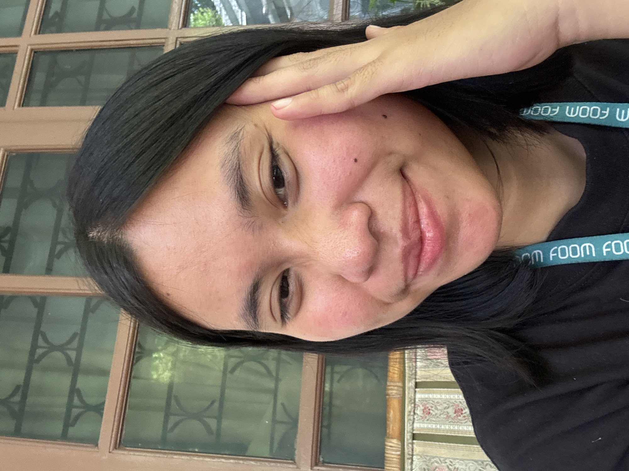 The image shows a close-up, waist-up shot of a young woman with dark hair and fair skin. She is looking towards the camera with a slight smile and her right hand is raised to her temple. She is wearing a black t-shirt with a teal lanyard around her neck. The background appears to be a window with wooden panes and a patterned upholstery on a couch. The image was taken outdoors, suggesting a residential or public space. The lighting is natural and appears to be from the side. The overall setting suggests a casual, everyday moment.