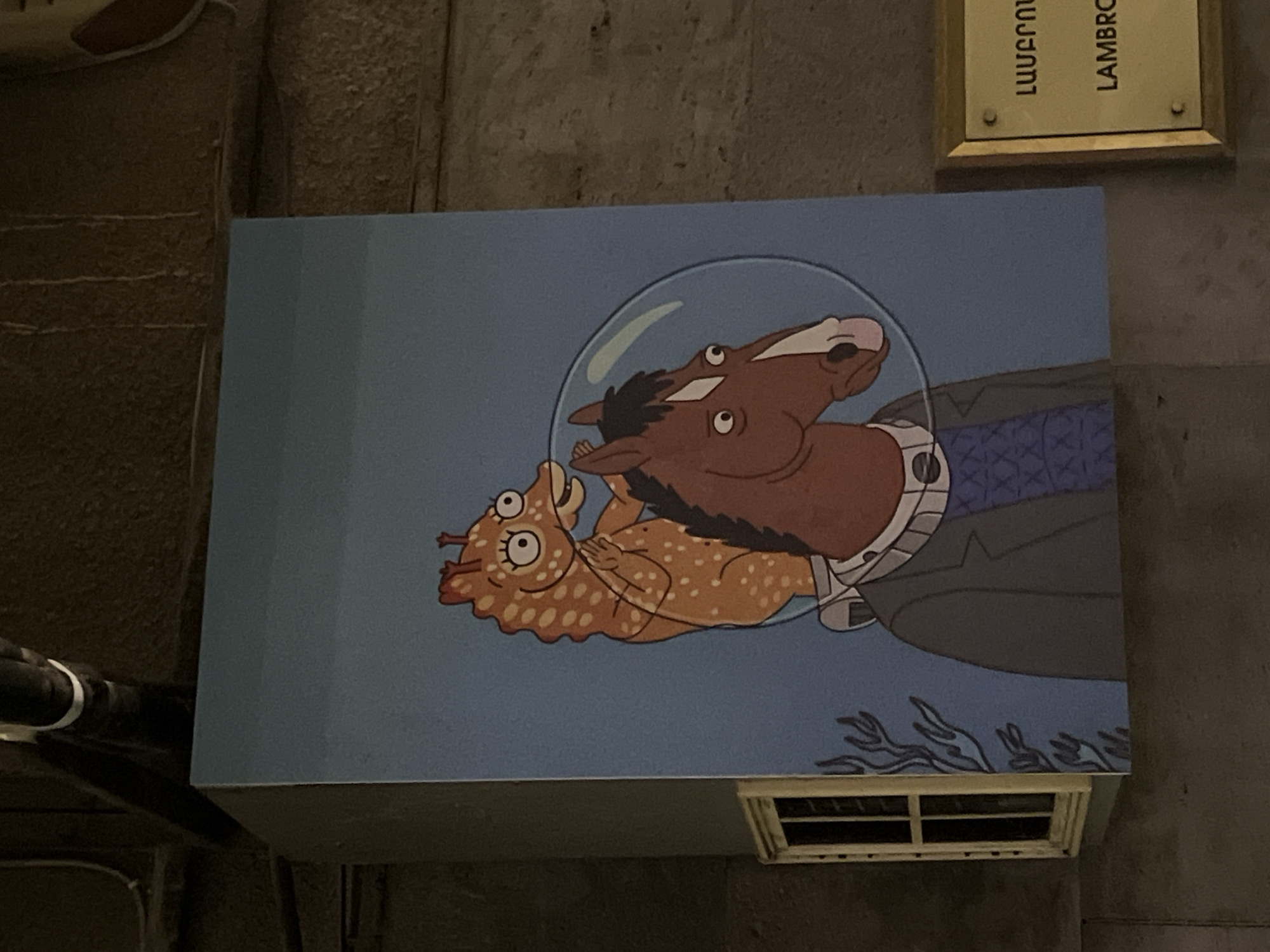 A rectangular panel affixed to a wall displays an image of BoJack Horseman, a cartoon horse in an astronaut's helmet, holding Diane Nguyen, a cartoon human character, on a blue background with abstract black wavy lines at the bottom. The panel appears to be mounted above a ventilation grille. A sign with Armenian script and the word "LAMBRO" in Latin script is visible above the panel. The location is Yerevan, Armenia.