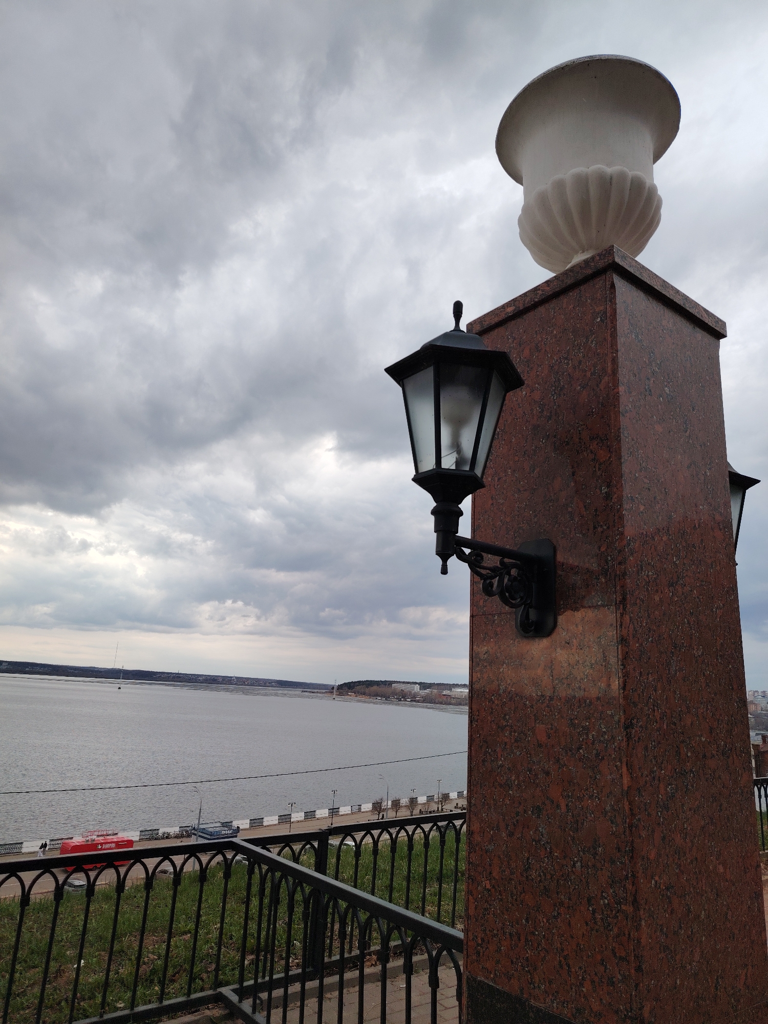 The image depicts a section of the embankment in Izhevsk, Russia, under a cloudy sky. In the foreground, a dark reddish-brown marble pillar is prominent, topped with a white decorative urn. An ornate black lamppost is attached to the side of the pillar. A black wrought-iron fence runs along the embankment, separating the viewer from the waterfront. The waterfront features a paved walkway with benches and streetlights. A red vehicle is visible in the distance. Across the water, the opposite bank features a cityscape, with buildings and a bridge visible. The overall scene conveys a somewhat somber or overcast atmosphere due to the cloudy sky and muted colors.