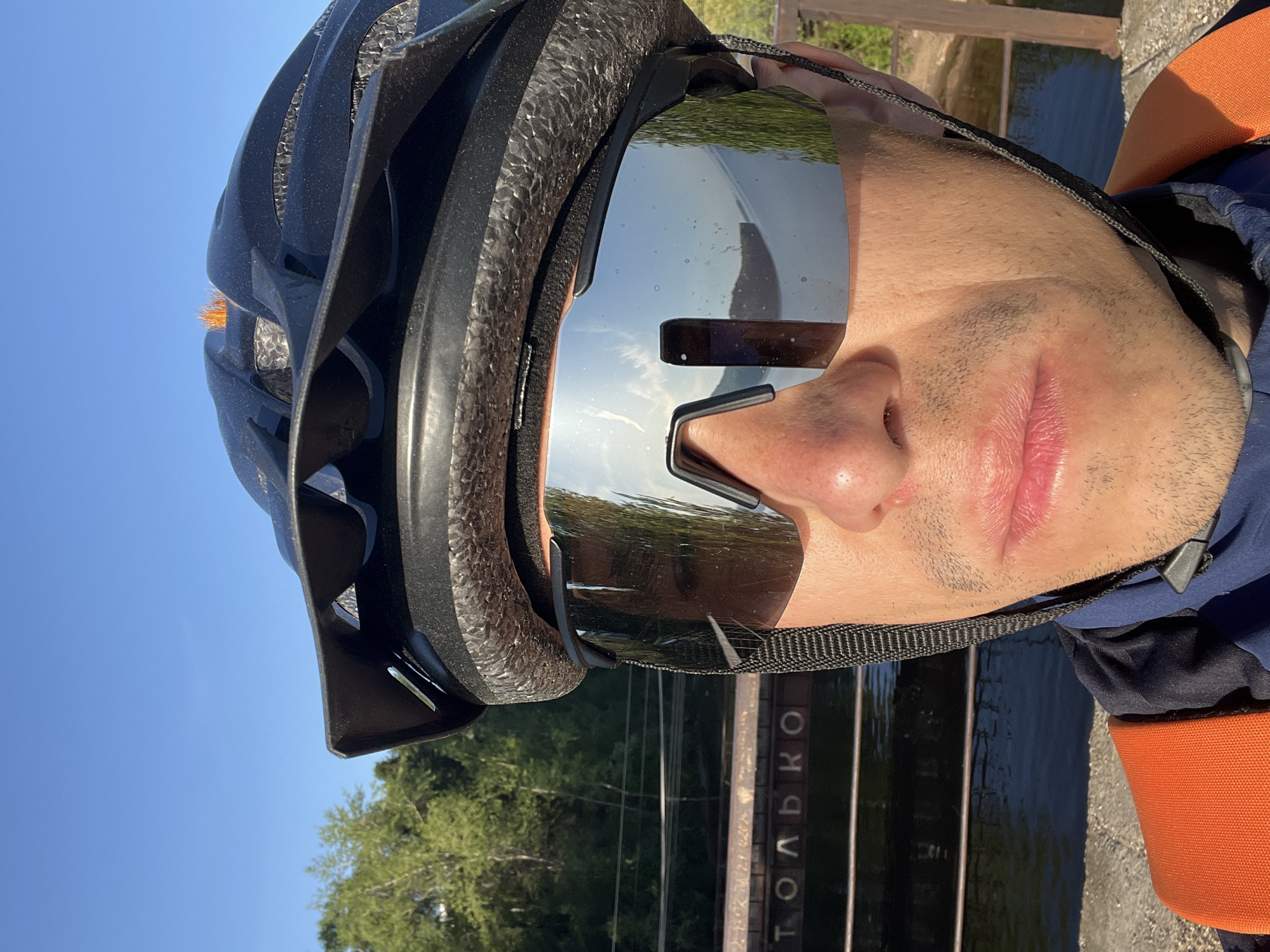 A person wearing a black cycling helmet and mirrored sunglasses is pictured outdoors in Chaikovsky, Russia. The scene includes a body of water and a metal structure with Russian Cyrillic lettering. The person appears to be near a bridge or embankment. The environment suggests a sunny day with visible trees and sky. The person is wearing clothing with orange and blue accents.