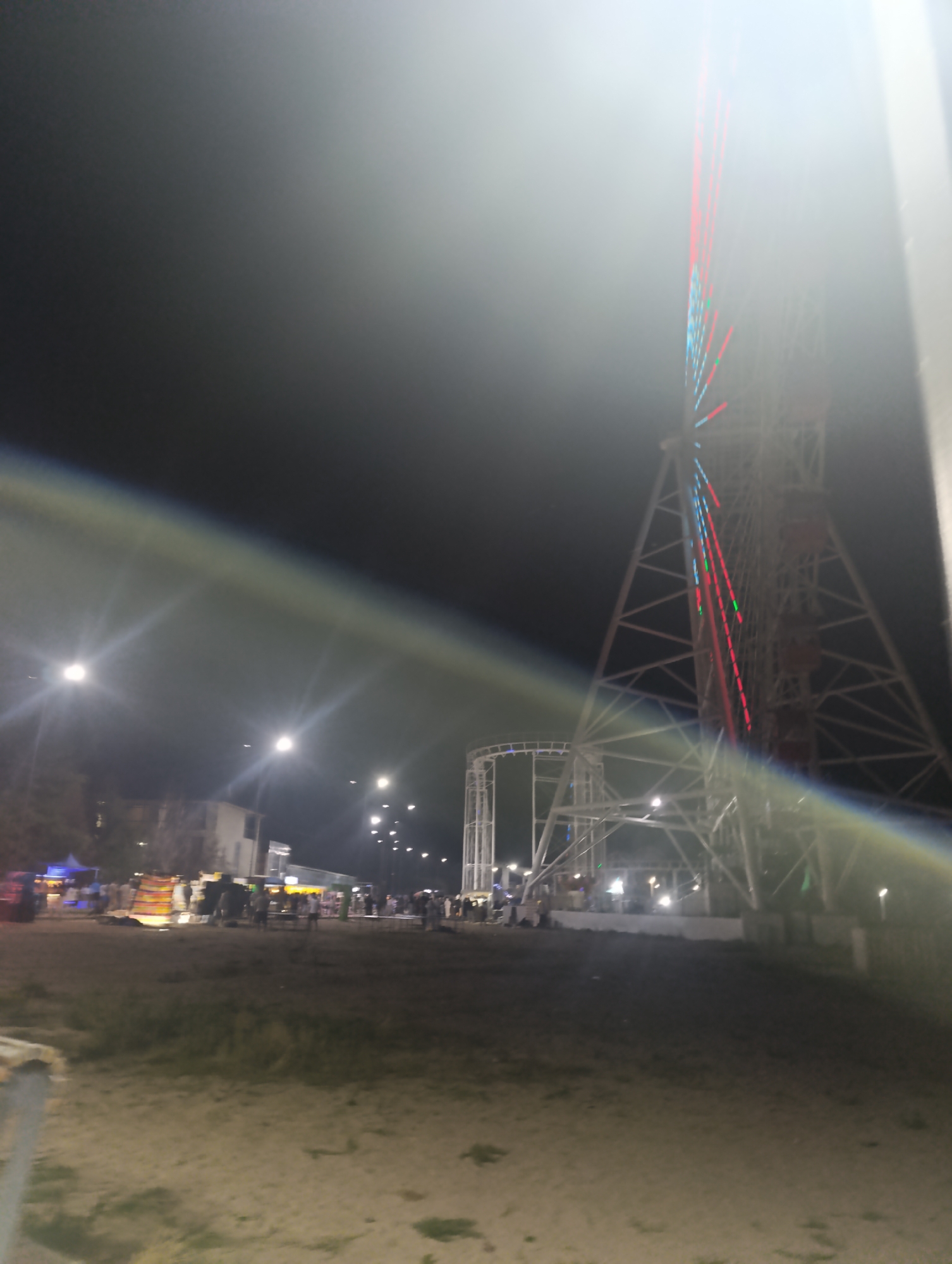 The image depicts a night scene at what appears to be an amusement park or fairground in Bosteri, Kyrgyzstan. A large Ferris wheel, illuminated with red and blue lights, dominates the right side of the frame. To the left, a roller coaster structure is partially visible. In the middle ground, there are several stalls or booths, suggesting commercial activity. A group of people, appearing as silhouettes, are gathered around these stalls. Streetlights illuminate the sandy ground, which is uneven and sparsely vegetated. The sky is dark, indicating it is nighttime. A lens flare streaks across the upper portion of the image, affecting the visibility of some details.
