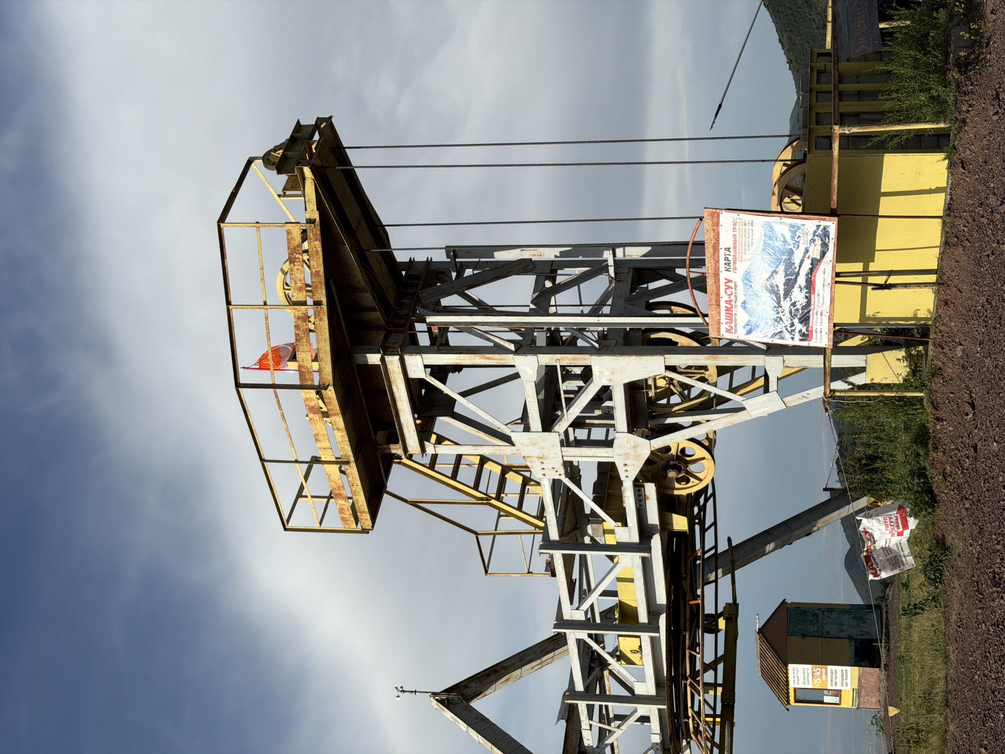 The image depicts an outdoor scene at a ski resort in Kashka Suu, Kyrgyzstan. In the foreground, a yellow structure supports a complex metal construction, likely part of a ski lift or cable car system. Several thick cables hang from the structure, disappearing out of frame. A large, colorful map of the resort's mountain tracks, titled "KAШKA-CCY KAPTА" and "ГОРНОЛЫЖНЫХ ТРАС," is prominently displayed on an easel to the right. To the left, a small, colorful booth with a window and a door stands near the ski lift structure. A weathered sign is visible on the side of the booth. In the background, rolling hills and mountains under a cloudy sky suggest a mountainous landscape. The ground in the foreground is covered in dark, rocky soil and some patches of dry grass. A few pieces of white debris, possibly posters or signs, are scattered on the ground near the map. No people are visible in the image. The overall impression is that of a functional but somewhat aged ski resort facility.