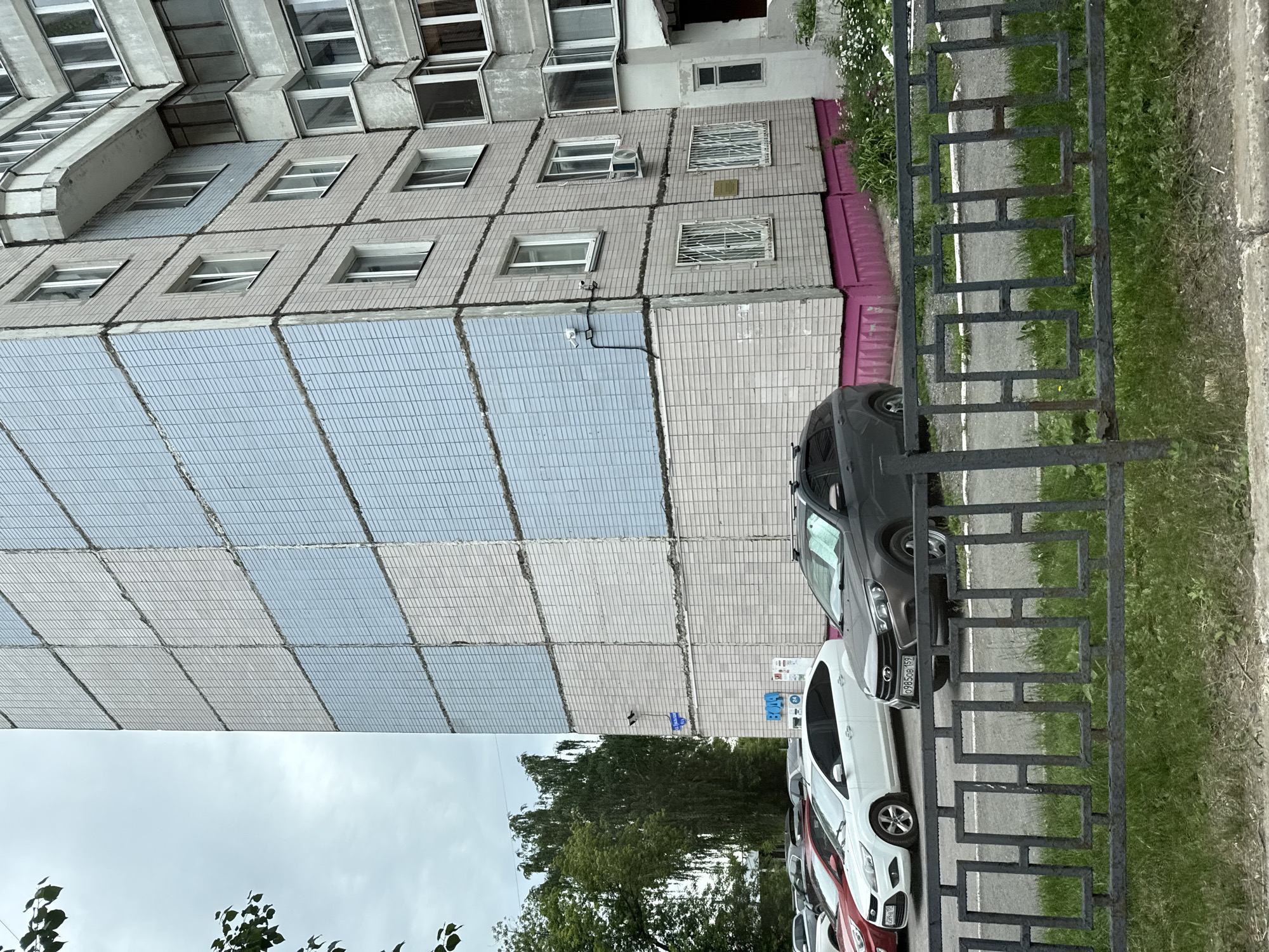 The image shows a multi-story residential building with a facade covered in light pink and light blue tiles. The building has a concrete balcony and several windows, some of which are open. There are two security cameras mounted on the side of the building. Below the building, there is a parking lot with several cars, including a white sedan and a dark gray hatchback. A black metal fence runs in front of the parking lot. Green grass and trees are visible in the background. The scene is set in the city of Perm, Russia, with a street sign visible on the building. The sky is overcast.