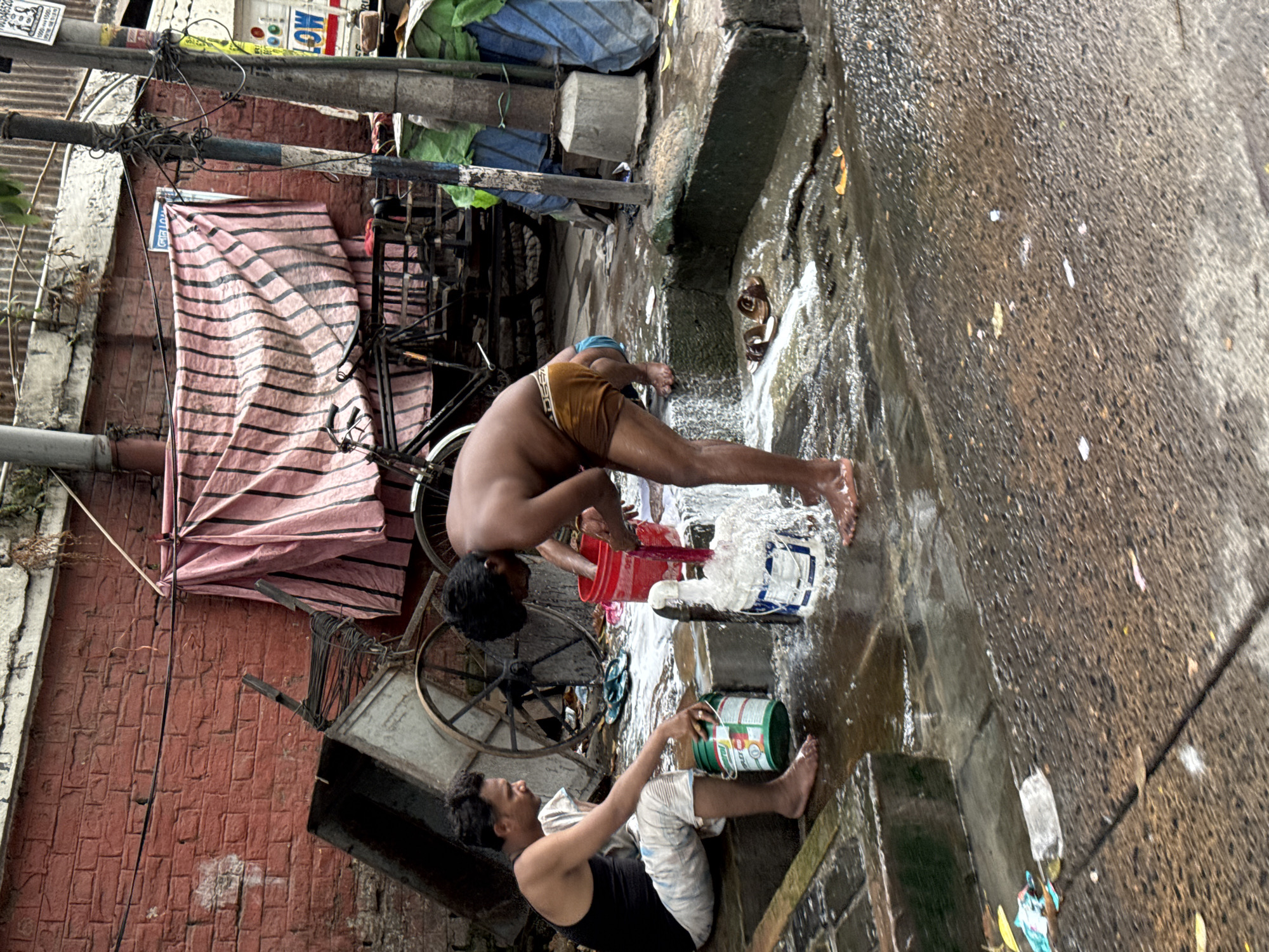The image, rotated 90 degrees clockwise, depicts two men bathing on a wet, concrete surface in an urban setting, likely an alleyway or street in Kolkata, India. The ground is slick with water and suds, suggesting the men are using soap.
In the foreground, a man wearing a dark tank top and light shorts sits on the ground, holding a green and white bucket. He appears to be washing himself, with water flowing from the bucket. Behind him, a second man, shirtless and wearing brown underwear, stands bent over, pouring water over his back from a red bucket. Both men are barefoot and focused on their activity.
The background features a rustic brick wall to the left, partially covered by a pink and black striped tarp. A bicycle and a handcart or rickshaw frame are visible leaning against the wall. Overhead, a tangle of utility poles and wires crisscross the frame, indicating a densely built-up area.
Notable details include a blue sign attached to the striped tarp with Bengali text, reading "জল না খাওয়ার" (Jol na khaowar), which translates to "Do not drink water" or "Not for drinking water." Another smaller, faded sign in the upper left corner has partial English text, "WATER SUPPLY" and a phone number. The brand "TRISHUL" is partially visible on one of the buckets. The scene captures a mundane but essential daily activity in a bustling, utilitarian urban environment under what appears to be diffused daylight.