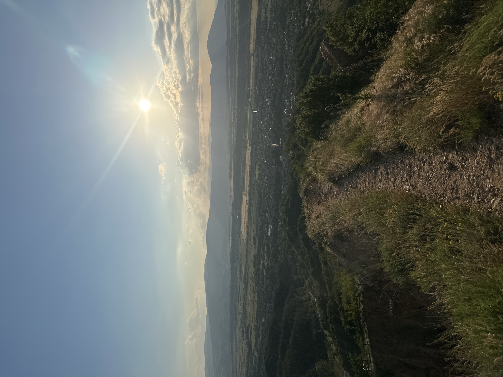 The image depicts a panoramic landscape view captured from a high elevation in the vicinity of Stob, Bulgaria. The time of day is characterized by a low-angle sun in the upper right quadrant, producing distinct lens flares and sunbeams. The sky transitions from light blue overhead to a warmer, lighter hue near the horizon, featuring scattered white and grey clouds, some partially illuminated by the sun.
In the immediate foreground, a narrow, unpaved dirt path, approximately 1-1.5 meters wide, traverses a ridge. The path is flanked by tall, dry, golden-brown grasses and some greener foliage, and it curves subtly towards the right of the frame. To the left of the path, a steep, eroded slope descends sharply. A section of a light-colored wooden fence or railing is partially visible on a lower ledge on the left, indicating a potential overlook or managed area. No human figures, animals, or specific objects are discernible on the path or the immediate foreground.
The midground displays an expansive valley system. Below the foreground ridge, densely forested slopes transition into a broad, relatively flat agricultural plain. This plain is composed of a mosaic of cultivated fields, appearing in varying shades of green and yellowish-brown. A narrow, serpentine body of water, likely a river or stream, is visible cutting across the plain from left to right.
The background features a range of hazy, undulating mountains forming the distant horizon. Below these mountains, a settlement, consistent with the description of Stob village, is visible within the valley. It comprises clusters of buildings and what appear to be roads, surrounded by a dense canopy of trees. The overall atmosphere indicates clear weather with some natural atmospheric haze.
