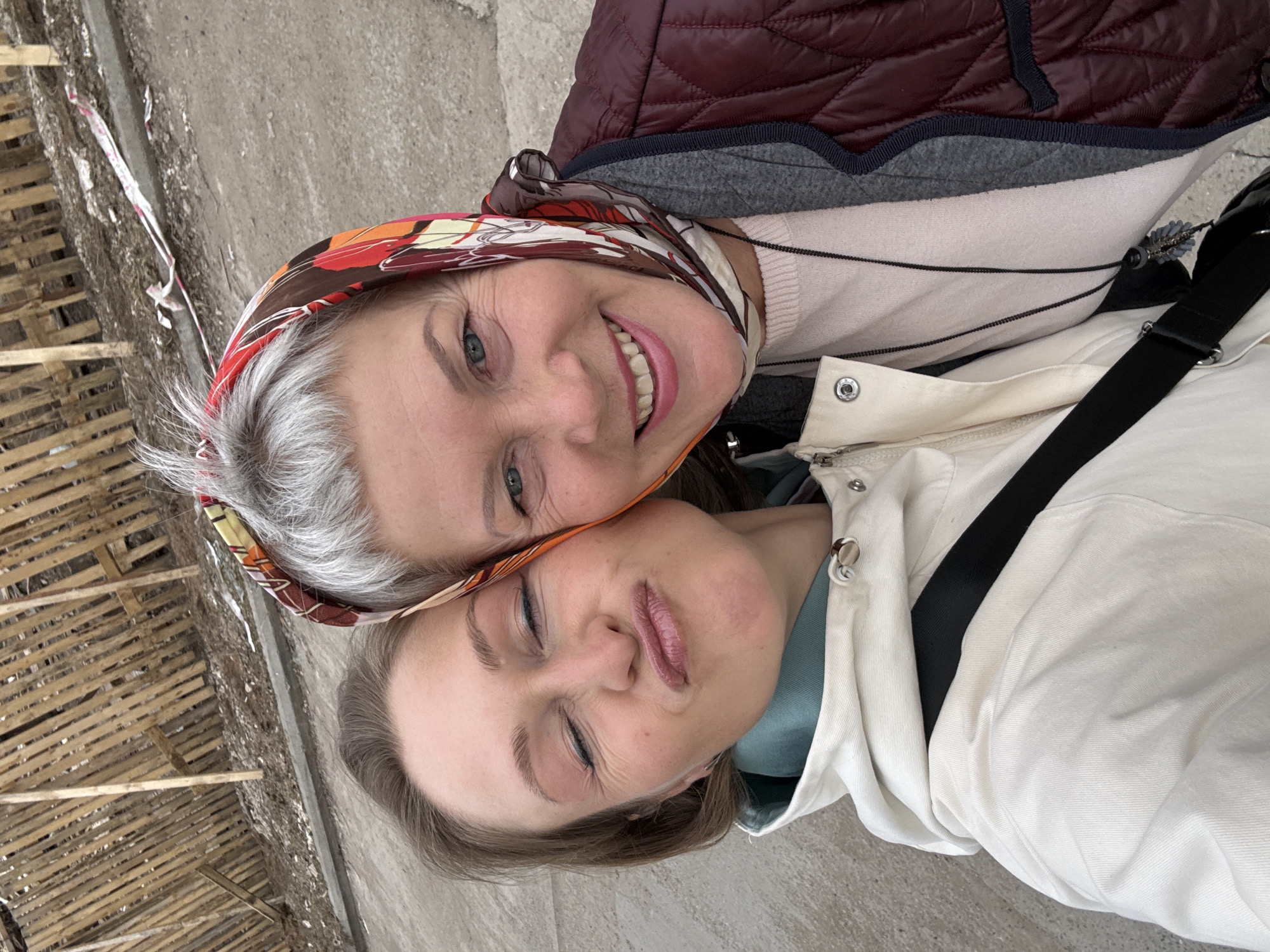 Two women are posing closely together for a photograph. The woman on top is older, with gray hair and a colorful headscarf, and is smiling broadly. The woman on the bottom is younger, with her eyes closed and a somewhat pursed expression. The women are outdoors, with a building structure partially visible in the background on the left side of the image. The ground appears to be paved. Based on the information provided the location is in the city of Chaikovsky, Russia.