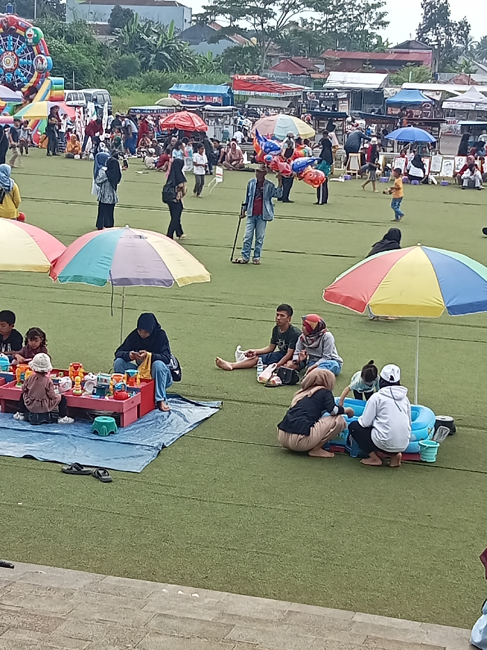 This image captures a vibrant outdoor gathering or market event taking place on a wide green artificial turf field in Singaparna, Indonesia. The scene is bustling with numerous people, including adults and children, engaged in various leisure activities under an overcast daytime sky.
In the foreground, several small groups are settled on the turf. To the left, a woman wearing a hijab is seated beside a red plastic play table, where a young child is engrossed in playing with colorful toys, including what appears to be a tea set. A blue tarp is spread beneath them, and sandals rest nearby. Further to the right, another group of individuals, including a man and a woman, are seated on the ground, seemingly relaxing. Next to them, a woman and a child are crouching around a small blue inflatable pool or basin filled with water, suggesting playful interaction. Colorful, striped beach umbrellas are positioned throughout the foreground, providing shade.
The middle ground features more people walking around, including a man with a cane carrying a bunch of colorful inflatable balloons, likely a vendor. Many women are dressed in hijabs, reflecting the local culture. In the background, a large number of market stalls or food vendors are visible, set up under various canopies. These stalls suggest a range of goods or food items being sold. A large, colorful inflatable structure, resembling a Ferris wheel or bounce house, stands prominently on the far left, indicating family-friendly entertainment. Beyond the field, residential buildings and lush trees line the horizon under a soft, diffused light, suggesting pleasant weather without harsh sunlight. The overall atmosphere appears relaxed and communal, with people enjoying a day out. No specific text is clearly legible on any signs or objects.