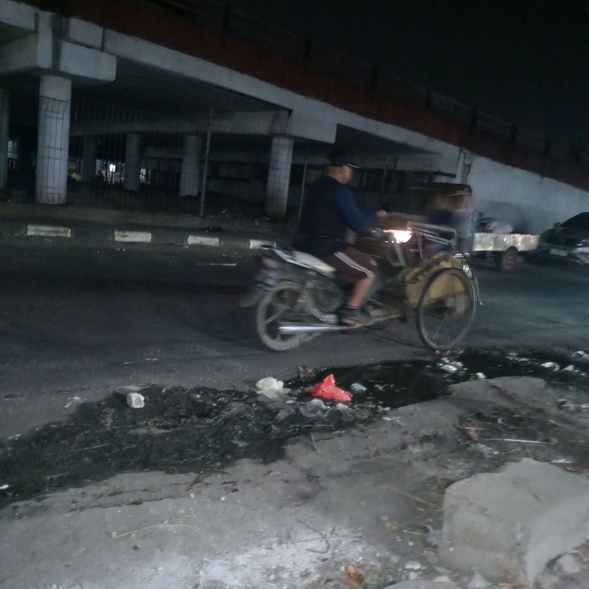 A person is riding a three-wheeled motorcycle on a paved road at night under an elevated concrete structure in Cileungsi, Indonesia. The motorcycle is dark colored with a white seat and has a single rear wheel. The rider is wearing a dark long-sleeved shirt and dark shorts with white stripes. A trailer with a silver box is attached to the back of the motorcycle. Further down the road, a dark car is parked. The road surface is dark and appears wet in places, with debris scattered along the edges, including white plastic bags and red plastic bags. The elevated structure is supported by large concrete pillars. The scene is dimly lit, with streetlights and lights from the motorcycle illuminating the area.
