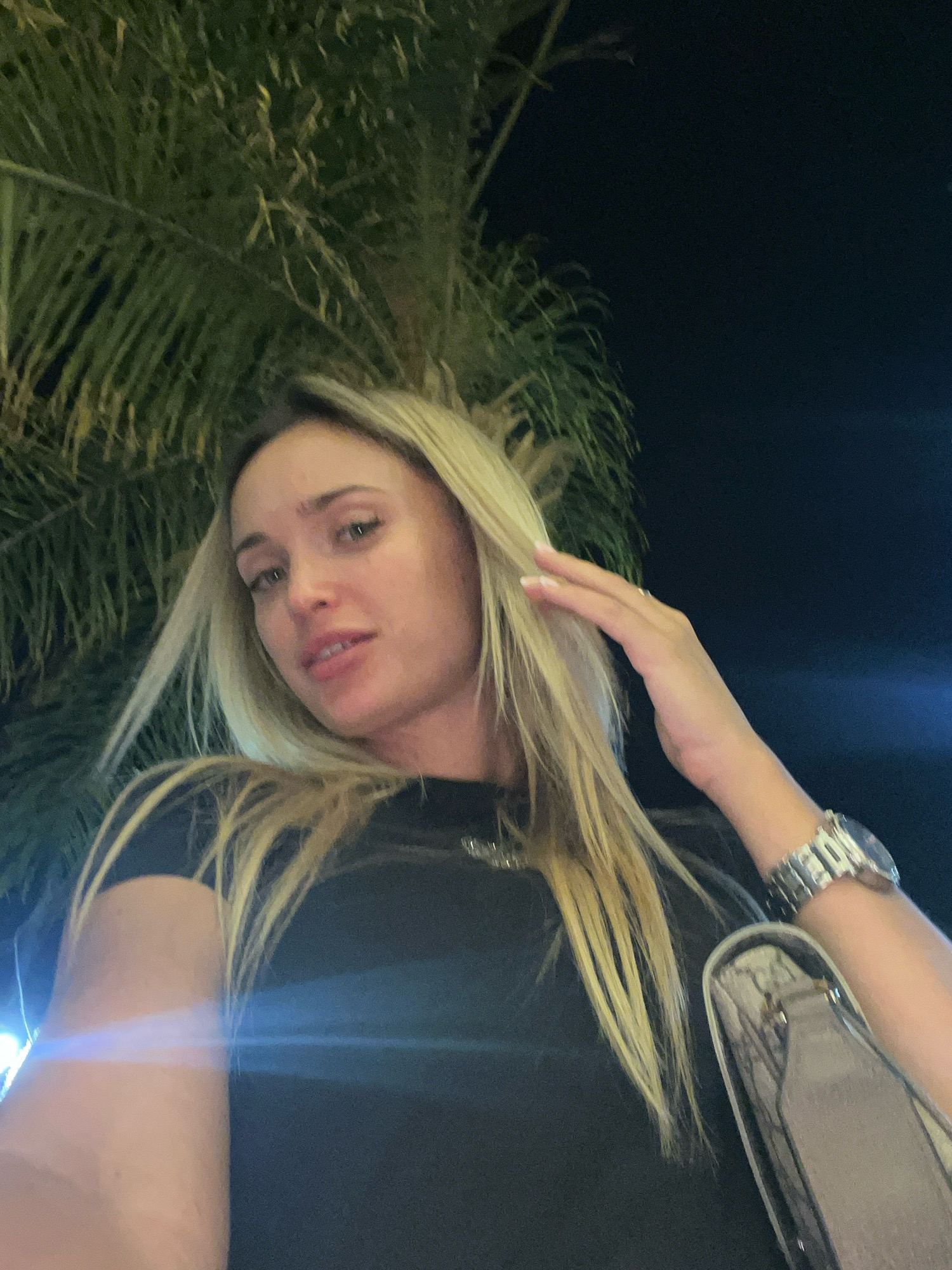 A close-up, low-angle, outdoor selfie shows a fair-skinned woman with blonde hair. She is wearing a black t-shirt and a large silver watch. Her left hand is touching her hair. The background is dark, with out-of-focus palm fronds in the upper left and a bright blue light flare across the right side. The scene appears to be at night, likely in Sharm El Sheikh, Egypt, given the tropical foliage and bright lights.