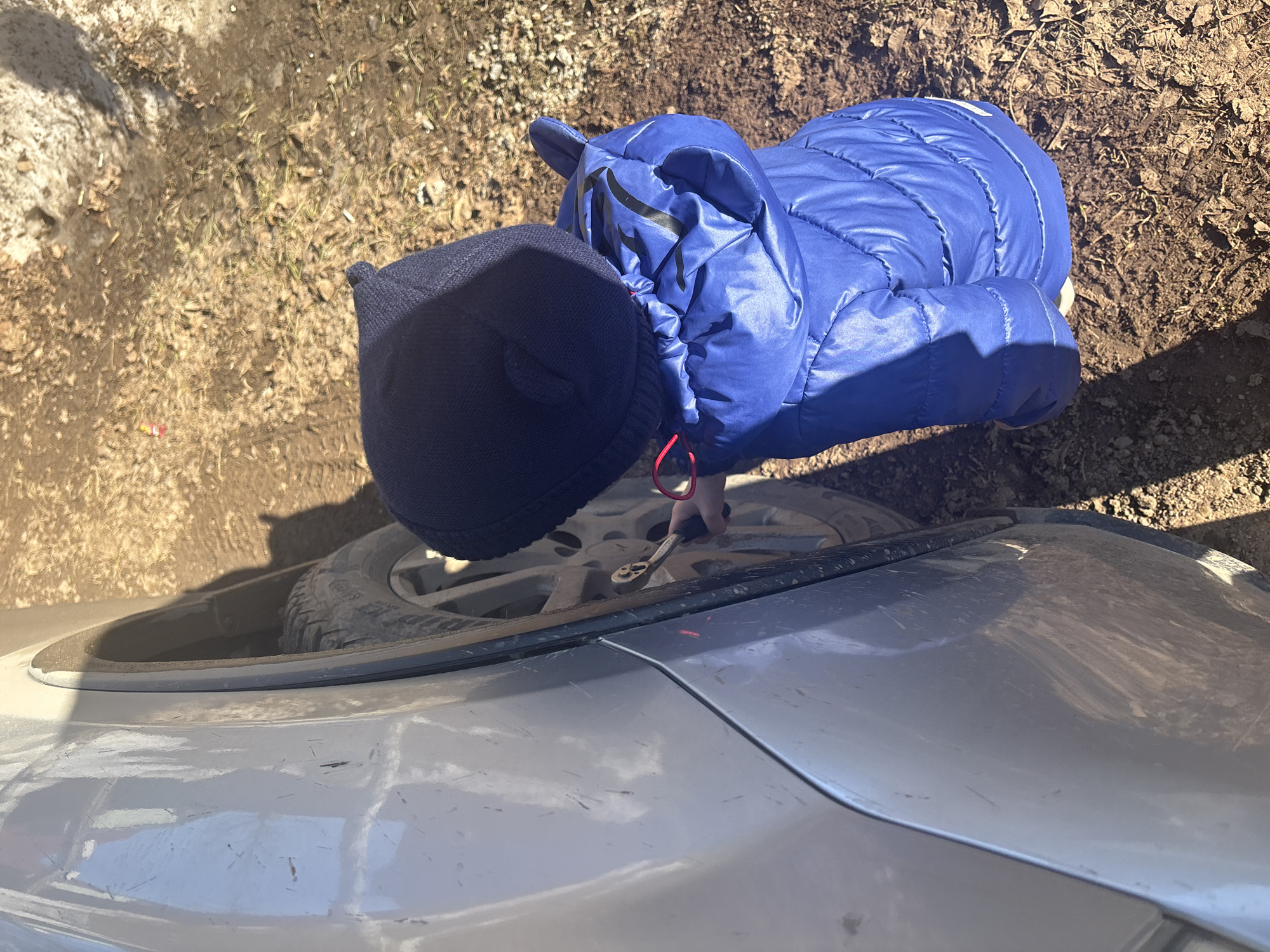A person wearing a dark blue knit cap and a shiny blue puffy jacket is using a wrench to work on the wheel of a silver car. The car is parked on a patch of dirt and leaf litter. The scene is likely outdoors. The specified city is Ферма, Russia.