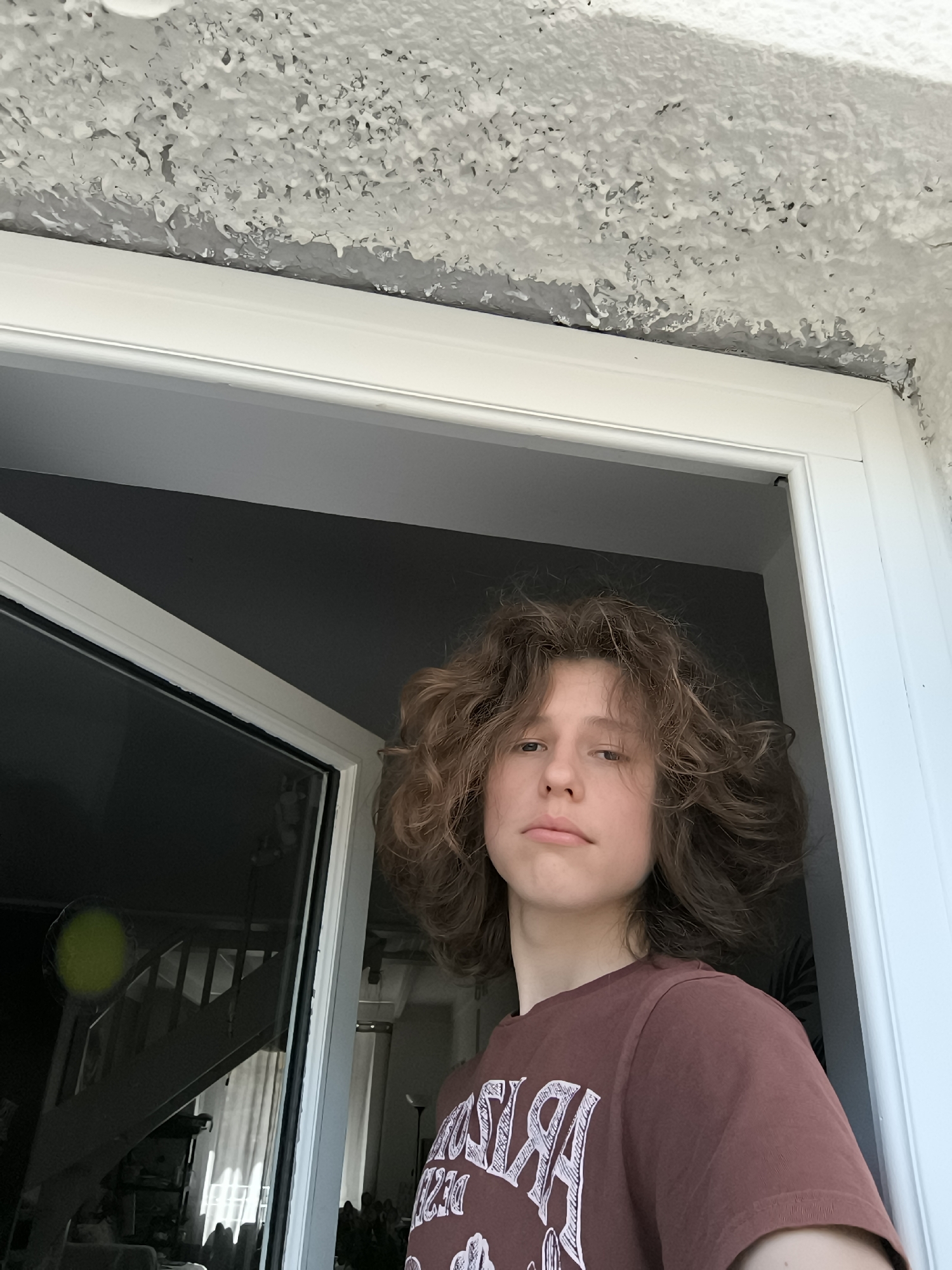 A person with curly brown hair and a brown t-shirt stands in a doorway. The person is centered in the frame and looking at the camera with a neutral expression. The doorway is white and appears to lead into a room. Through the doorway, a staircase and other interior elements are visible, reflected in the glass window. The exterior of the doorway reveals a textured white ceiling or wall. The location is impossible to discern from the image without additional context. Pélussin, France cannot be determined.