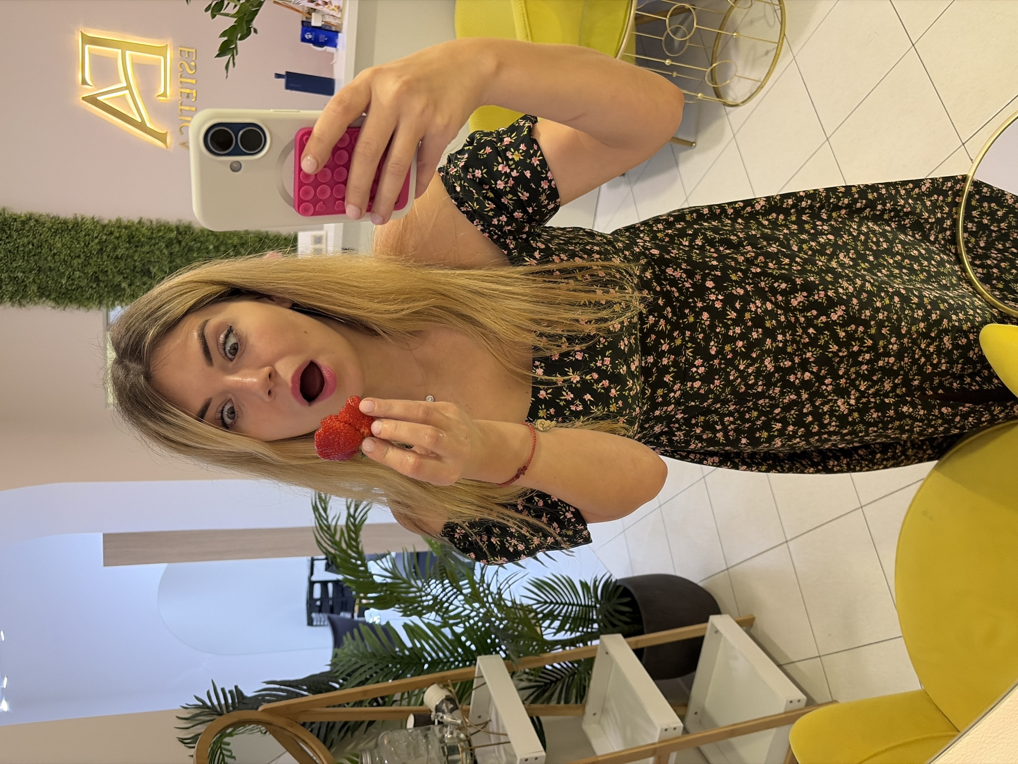 The image shows a young woman with blonde hair, holding a strawberry up to her mouth, with an astonished expression on her face. She is taking a selfie with a smartphone. She is wearing a black floral dress with off-the-shoulder sleeves. In the background, there is a potted plant, a shelf with items, and a large illuminated sign that reads "EA ESTETICA." The floor is tiled, and there is a yellow chair visible. The setting appears to be a beauty salon or spa, potentially located in Budva, Montenegro, given the context of other similar images.
