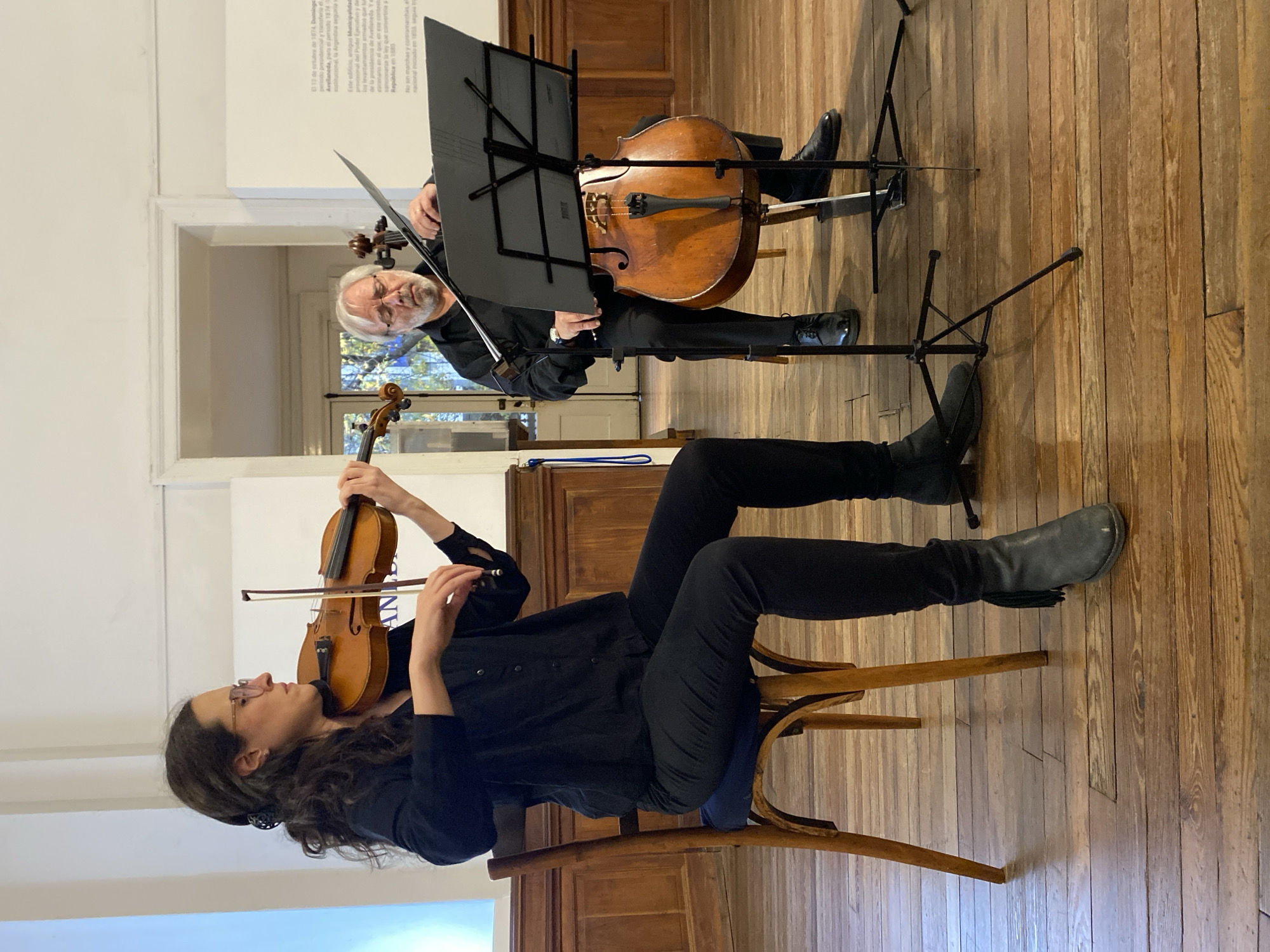 The image shows a musician playing a violin while seated on a wooden chair. She is wearing dark clothing and glasses. Another musician is standing behind her, playing a cello. Both have music stands in front of them. The setting appears to be indoors, possibly a historic building with hardwood floors. The text on the wall is in Spanish and references the city of Buenos Aires, Argentina, with references to the year 1874.