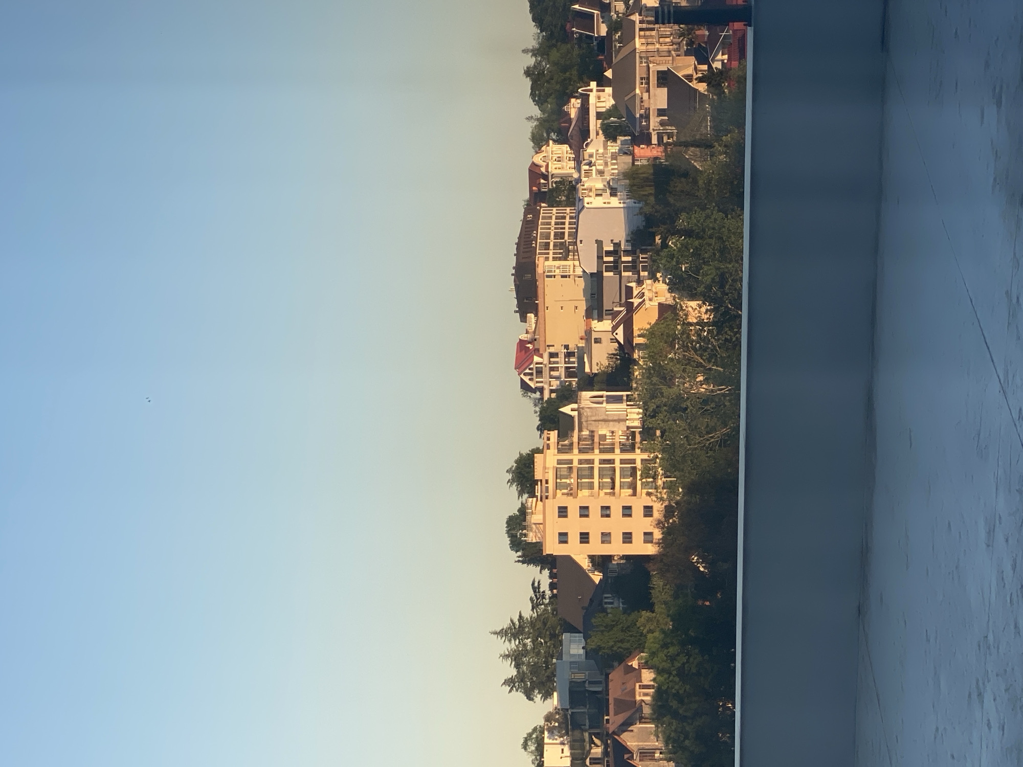 The image depicts a cityscape view taken from a high vantage point, possibly a balcony or window. The urban landscape consists of closely packed buildings, including residential houses and apartment complexes, interspersed with mature trees. The buildings exhibit diverse architectural styles and colors, ranging from light pastels to earthy tones. The sky occupies a significant portion of the image and presents a gradient of pale blue to light yellow, suggesting either sunrise or sunset. A structural element, perhaps a wall or railing, is visible in the lower right corner of the frame. Given the architectural style and densely built-up environment, it's consistent with a location like Sochi, Russia, although specific landmarks aren't identifiable.