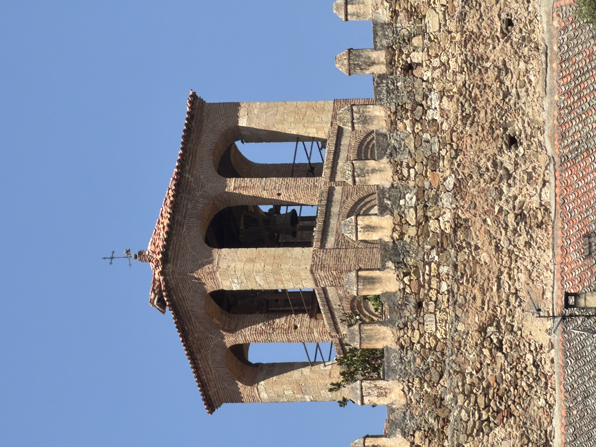 The image shows a bell tower constructed of brick and stone, situated atop a crenelated stone wall. The bell tower has an arched opening on each of its four sides, with bells visible through the openings on the left and right. A cross tops the structure. The wall is made of rough, irregular stones. In the foreground, the tiled roof of a building is visible. The scene is set against a clear blue sky. This is a view of the bell tower of the Church of San Andrés in Segovia, Spain.