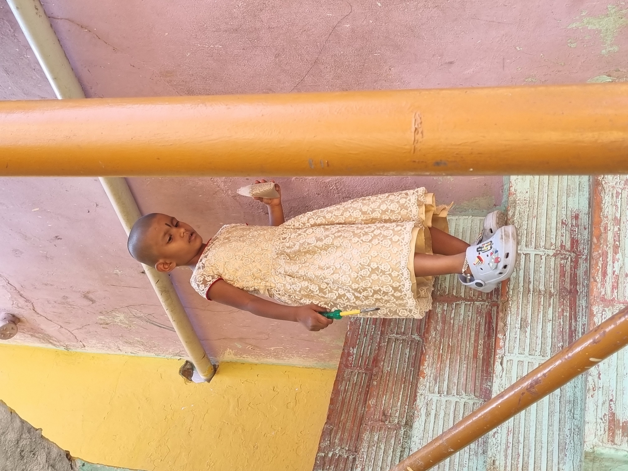 A young child with a shaved head and a red bindi on her forehead stands on a set of weathered stairs in Pedda Kammavaripalli, India. She is dressed in a light-colored, patterned lace dress with short sleeves and white Crocs-style sandals adorned with charms, and a silver anklet on her left ankle. The child holds a yellow and green screwdriver-like tool in her right hand and a small, clear plastic bottle containing a granular substance in her left hand. She is looking downwards and slightly to her left. The immediate environment includes a faded pink wall to her right and behind her, displaying cracks and texture. A bright yellow wall is visible to her left. A horizontal light-colored pipe runs along the pink wall near the top. A prominent vertical orange-brown pipe or pillar is positioned in the foreground on the right side of the image. A diagonal orange-brown pipe, likely a handrail, is visible in the lower left foreground. The stairs feature peeling light blue-green paint over a reddish-brown, textured brick or tile pattern.