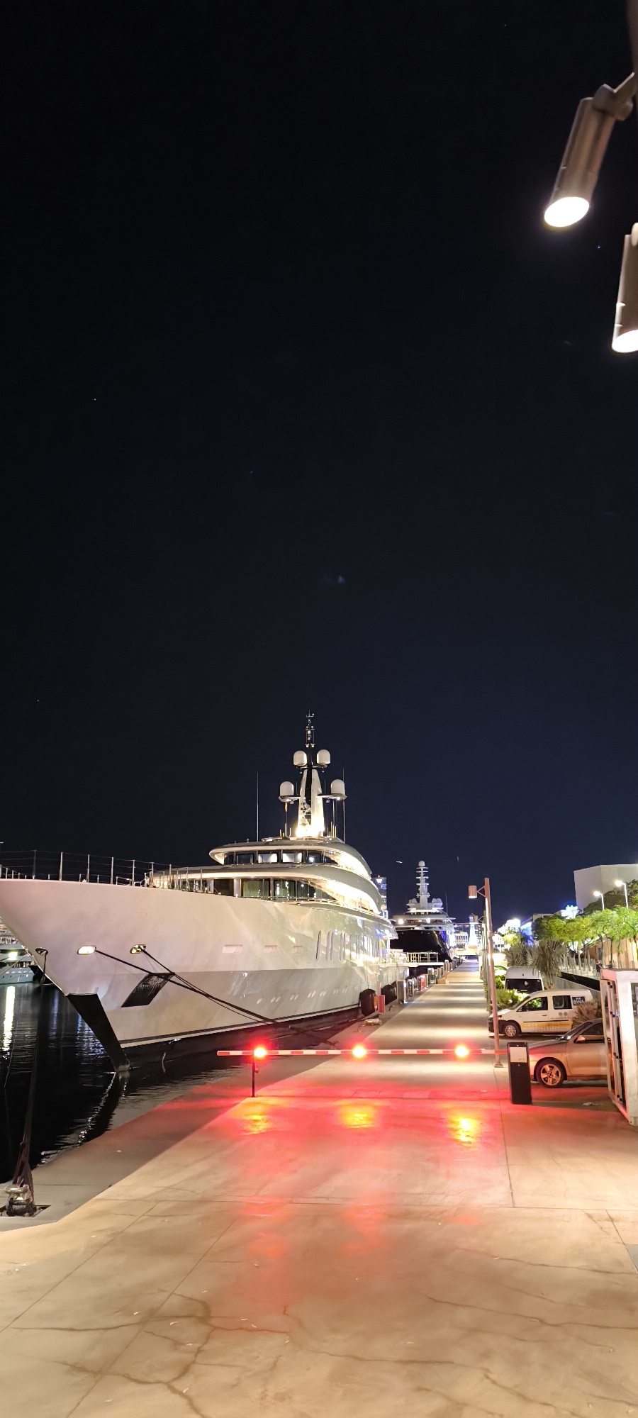 The image depicts a harbor scene at night, likely in Barcelona, Spain. A large, white luxury yacht is docked alongside a concrete pier. The yacht features multiple decks, windows, and a mast with communication equipment. Other yachts are docked nearby, but they are smaller and further in the background. The pier is illuminated by overhead lights and a red traffic barrier with lights. Several vehicles, including a white van and a silver car, are parked near the pier. The sky is dark, indicating nighttime. There are few people visible in the scene.