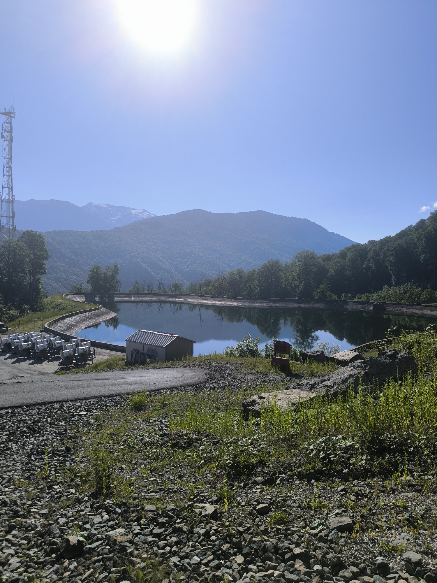 The image depicts an outdoor scene on a sunny day with a clear blue sky. In the foreground, there is a rocky, gravelly area with sparse green vegetation. A paved road curves through the scene. To the left, a cluster of white, cylindrical objects on trailers, possibly snow cannons or generators, are lined up. Adjacent to these is a rectangular building with a metal roof.
Further back, a large, artificial reservoir with a curved embankment is visible, filled with calm water that reflects the surrounding landscape and sky. The water body is bordered by green, forested hills that recede into the distance. In the far background, majestic mountains, some with snow-capped peaks, are silhouetted against the sky. A tall telecommunications tower stands on a hill to the left of the reservoir. The overall impression is a scenic mountain landscape with industrial elements. This location is likely within the Krasnaya Polyana ski resort area near Sochi, Russia, a region known for its mountainous terrain and winter sports facilities, where such reservoirs and equipment would be necessary for snowmaking. There are no people visible in the image.