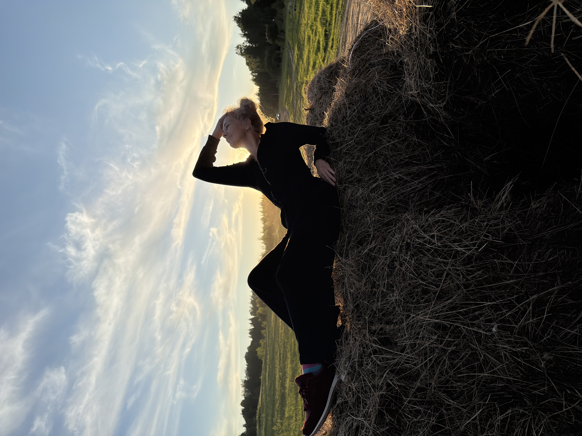 This vertically oriented image, rotated 90 degrees clockwise, captures a woman silhouetted against a brilliant sky during what appears to be the golden hour of either sunrise or sunset. The scene is set in an open, rural landscape, likely near Ozernoye-1, Russia.
The main subject is a woman, presented in profile, leaning against a substantial hay bale or mound of dry grass on the right side of the frame. Her head is turned to the left, and her left hand is raised to her forehead, shielding her eyes from the intense, low-angle sunlight that backlights her. Her hair, appearing light in color, is styled in an updo. She is dressed in dark, long-sleeved attire, possibly a jumpsuit, dark trousers, and reddish-brown athletic shoes with colorful striped socks (hints of blue and pink are visible). Her right hand gently rests on the textured surface of the hay.
The background features a vast expanse of sky, filled with soft, wispy clouds that glow with the warm, golden light of the sun. Below the horizon, a green field stretches into the distance, dotted with trees that also catch the warm light. The ground immediately around the woman and the hay bale consists of dry, natural terrain.
The activity suggests the woman is taking a moment to observe or enjoy the outdoor environment and the natural beauty of the light. Her pose conveys a sense of calm and contemplation. There is no visible text in the image.