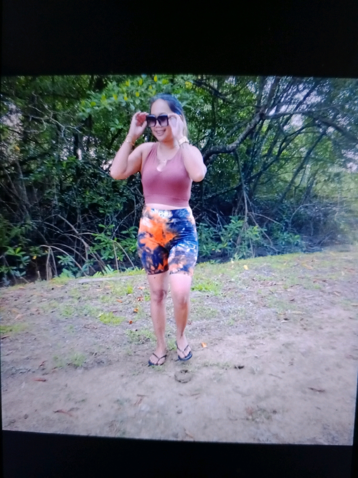 A full-body, medium shot captures a woman standing on a dirt path outdoors, with trees and foliage in the background. The woman is wearing a pink crop top, multi-colored tie-dye shorts, and black flip-flops. She has dark hair and is wearing large, square-shaped sunglasses, and gold bracelets on both wrists. She is smiling and adjusting her sunglasses with her right hand. The ground is a mixture of dirt and sparse grass, with some fallen leaves. The trees in the background are dense with green leaves and branches. The lighting suggests it is daytime.