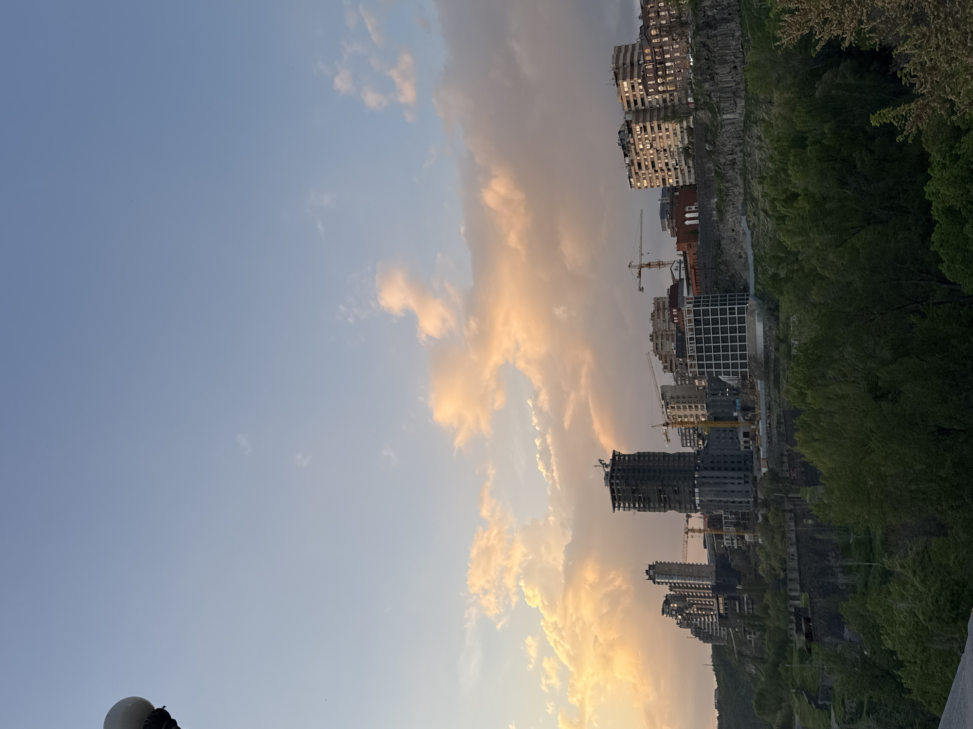 The image presents a vibrant urban landscape bathed in the warm light of either sunrise or sunset, viewed from a slightly elevated perspective. The sky dominates the upper portion, transitioning from a clear, light blue on the left to a dramatic display of soft golden-orange and pink clouds, indicative of the low sun's angle.
Below the colorful sky, a dense cityscape unfolds. Numerous buildings, varying in height and architectural style, populate the middle and lower sections of the frame. Some appear to be residential or commercial structures with visible windows, a few of which reflect the warm light. Interspersed among the completed buildings are several structures under construction, identifiable by their exposed frameworks and the prominent presence of multiple tall construction cranes. These cranes underscore ongoing development in the area.
To the right, a significant natural feature provides a stark contrast to the urban environment: a steep, rocky embankment or cliff face generously covered with lush green trees and foliage. This green belt suggests the city's integration with, or proximity to, natural terrain or a valley.
In the bottom-left corner, a small portion of a spherical, white object is visible, possibly part of a lamppost, monument, or railing. The overall lighting and cloud patterns suggest a peaceful yet dynamic moment in time. No discernible text is visible within the image. Given the context of Yerevan, Armenia, the scene captures a blend of urban expansion and natural beauty.