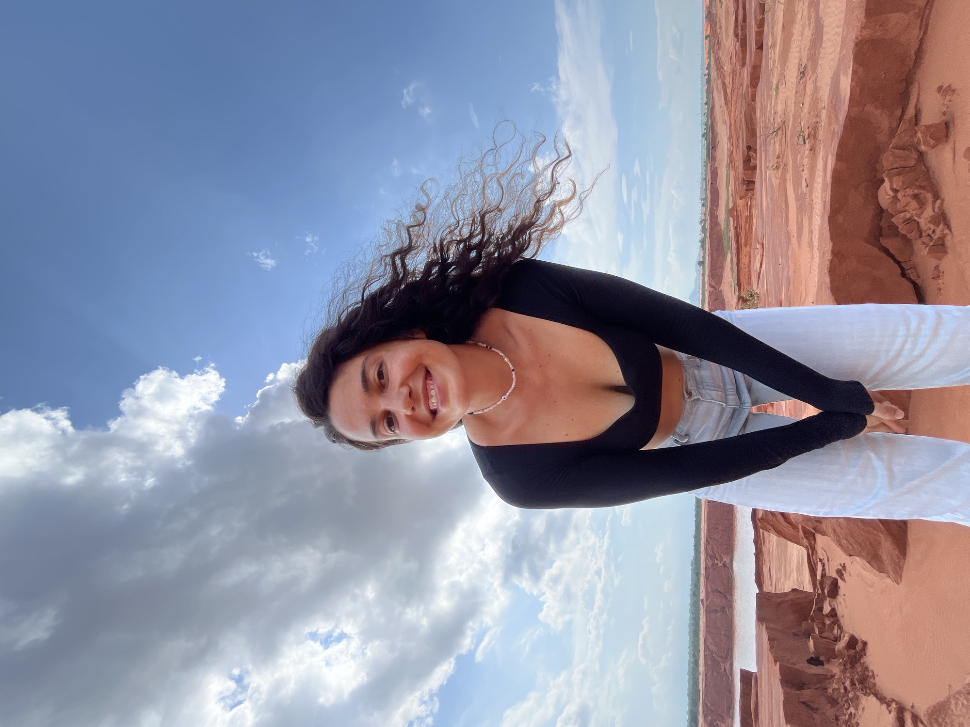 A woman with long, dark, curly hair is posing for a photo outdoors, her head tilted down, and her hands clasped together in front of her. She is wearing a black long-sleeved crop top, a pearl necklace, and light-colored pants. The background is a landscape of red sand dunes and a body of water, indicative of the sand dunes in Mui Ne, Vietnam. The sky is blue with scattered white clouds.