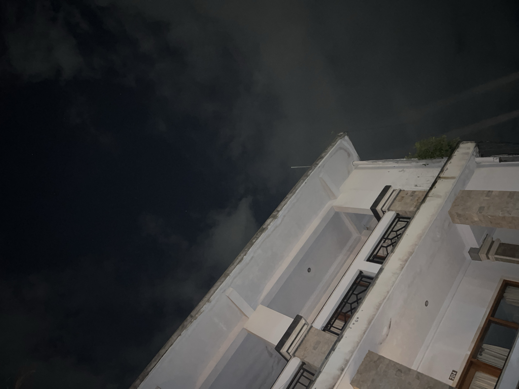 The image depicts an upward angled view of a white building against a dark, cloudy night sky. The building has multiple stories, with visible windows and balconies featuring dark metal railings. The structure appears to be located outdoors in Canggu, Indonesia. The presence of windows and balconies suggests a residential or commercial building. The number "204" is visible on the exterior of the building.