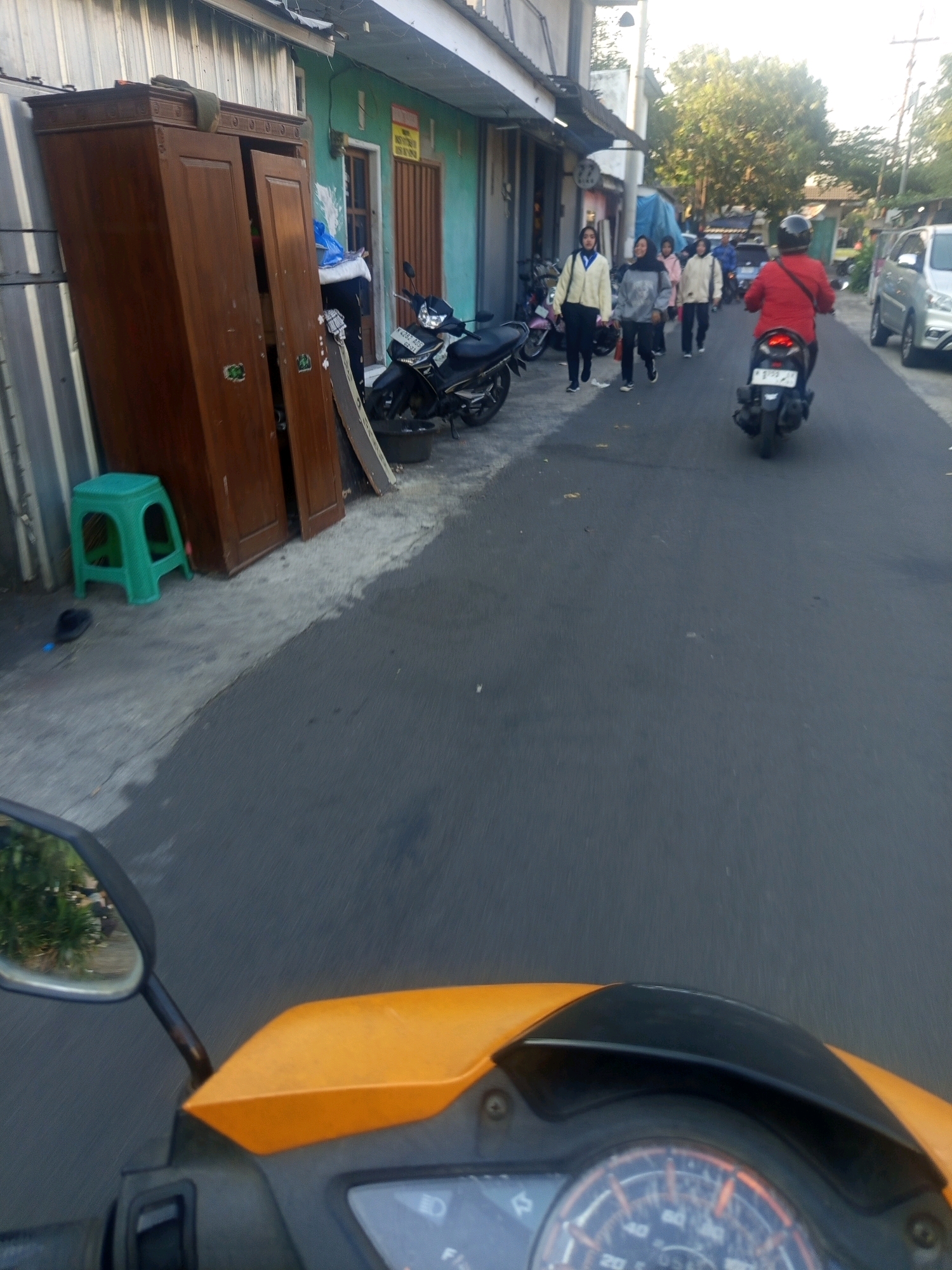 In an urban scene likely in Malang, Indonesia, a brown wooden wardrobe stands open on the left side of a paved road. A green plastic stool is situated near the wardrobe. Several people are walking on the right side of the road, with some riding motorcycles. One person in a red shirt is riding a motorcycle in the foreground, facing away from the camera. Further down the road, a silver car is visible. The environment appears to be a street with buildings lining one side and a road for traffic. The lighting suggests it is daytime, possibly late afternoon due to the shadows.