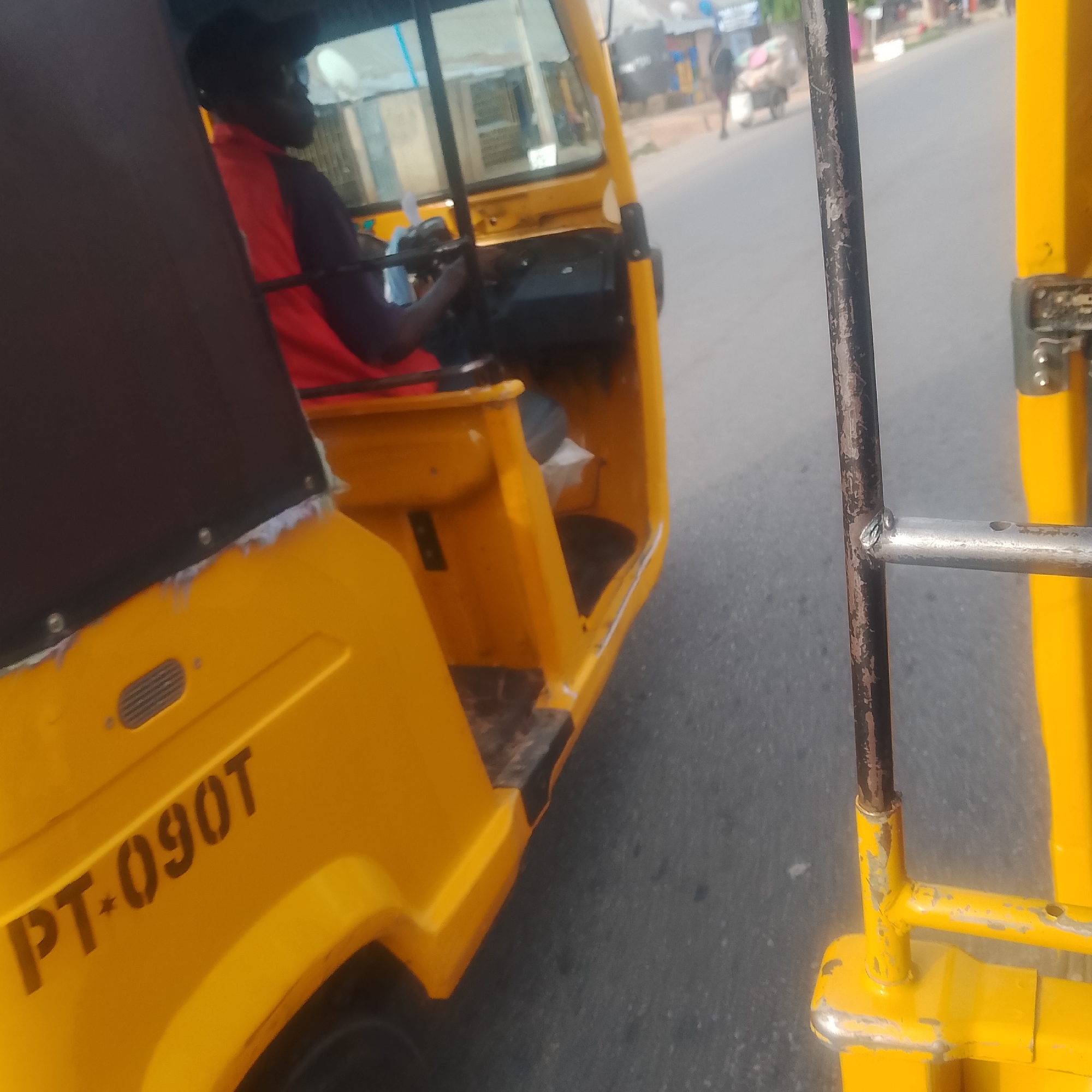 A driver wearing a red and blue shirt operates a yellow auto-rickshaw on a paved road in Jalingo, Nigeria. The auto-rickshaw is a three-wheeled vehicle with a covered passenger area and a driver's cockpit. The license plate number "PT-090T" is visible on the side of the vehicle. In the background, other vehicles and pedestrians can be seen, suggesting a typical urban environment. The overall scene depicts everyday life and transportation in Jalingo.