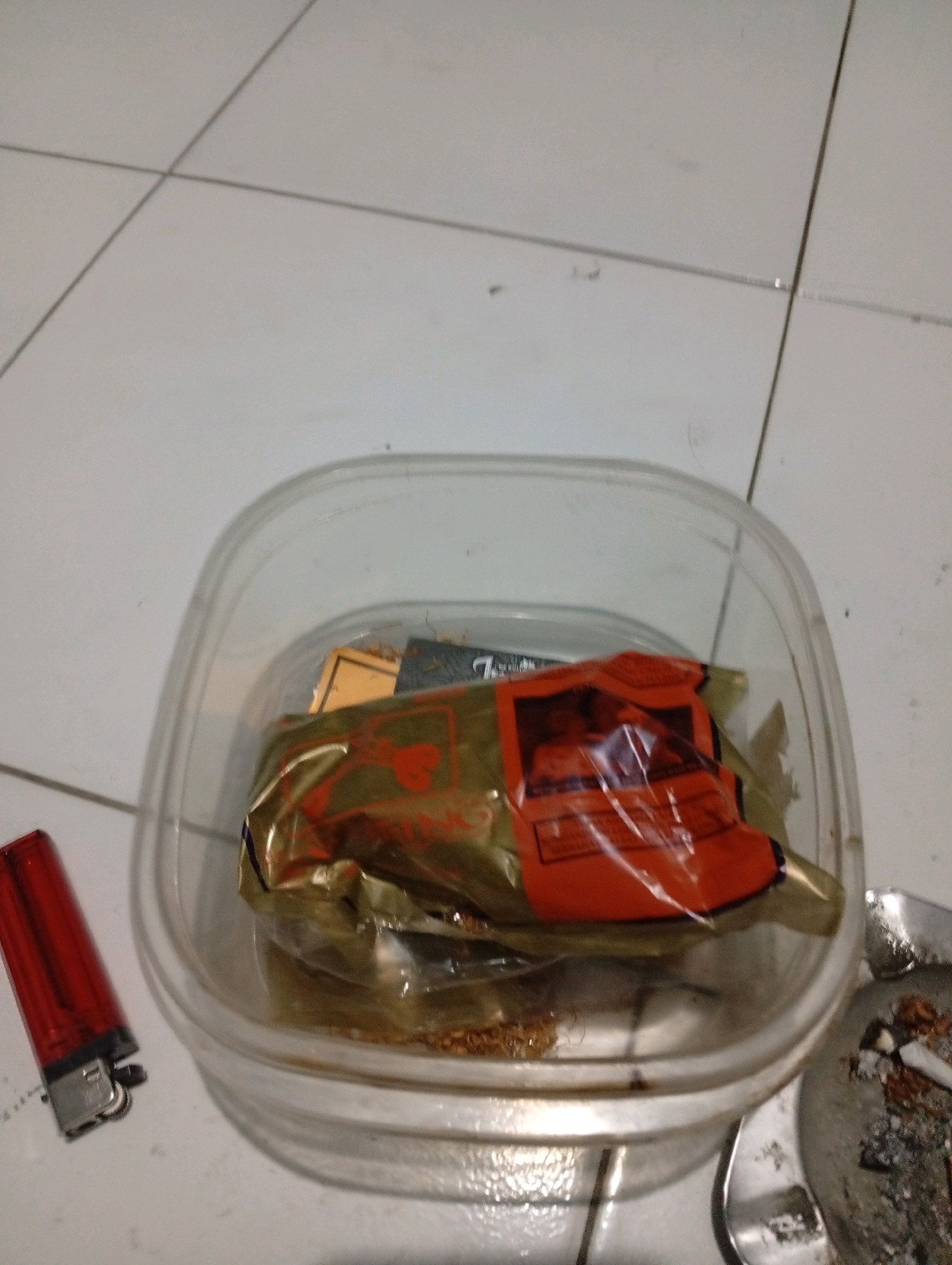 The image depicts a close-up, overhead view of a clear plastic container filled with loose tobacco. Inside the container, on top of the tobacco, is a crumpled, golden-colored pouch with an orange label featuring a red design. To the left of the container is a red disposable lighter. To the right of the container is a metal ashtray containing cigarette butts and ash. The scene appears to be indoors, possibly on a tiled floor, given the grout lines visible in the background. The lighting is dim, and the focus is on the objects within the container and their immediate surroundings. There are no people or animals present in the image, and no discernible geographical markers of Tasikmalaya, Indonesia, are visible.