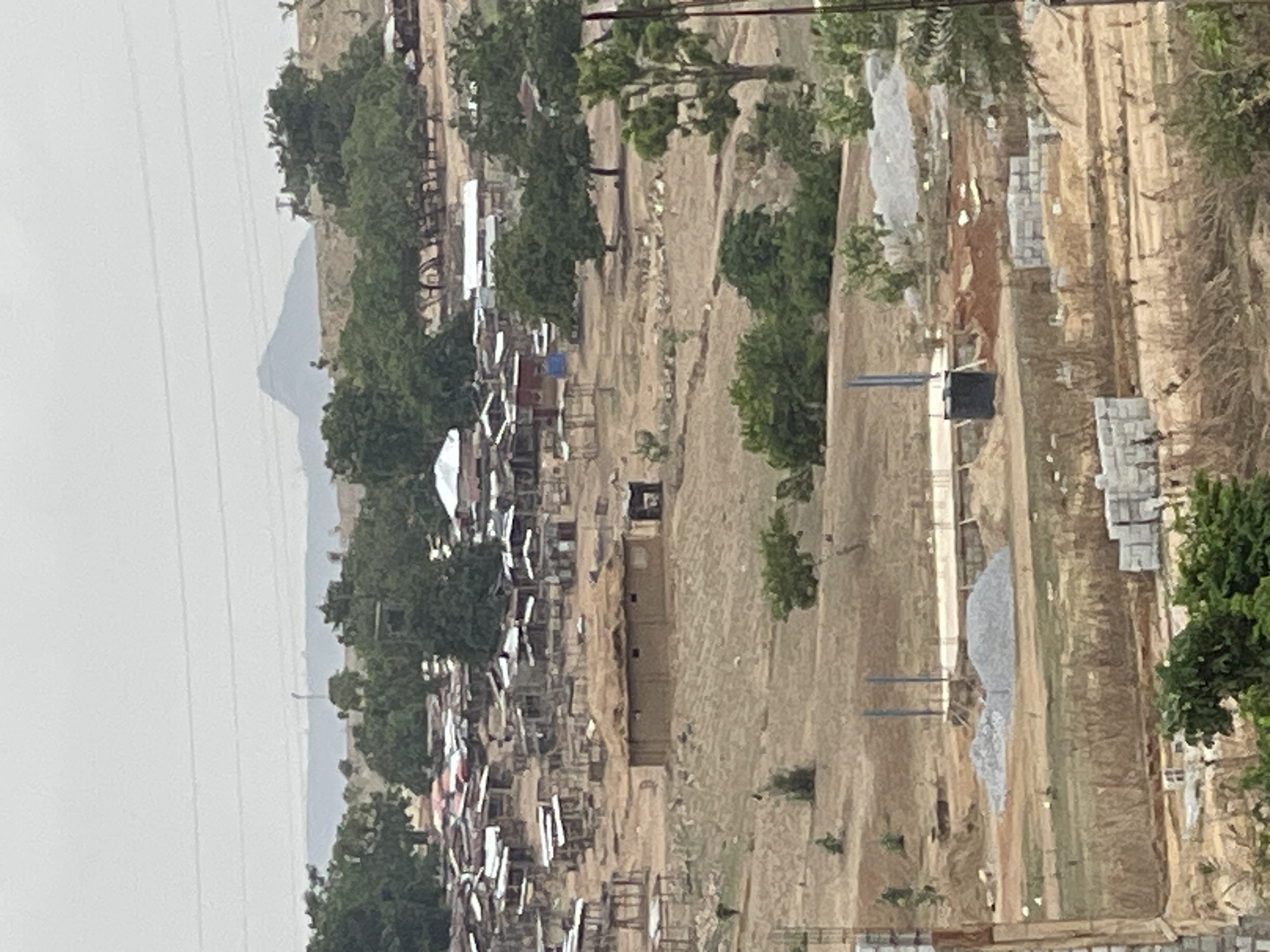 The image shows a view of Angwan Sayawa, Nigeria. In the distance, a mountain is visible against an overcast sky. In the foreground, there is a dense collection of buildings, many with corrugated metal roofs, interspersed with trees. Beyond this area, there appears to be a construction site with piles of materials and partially built structures. Vegetation is sparse, with dry, grassy areas. Overhead power lines are also visible.