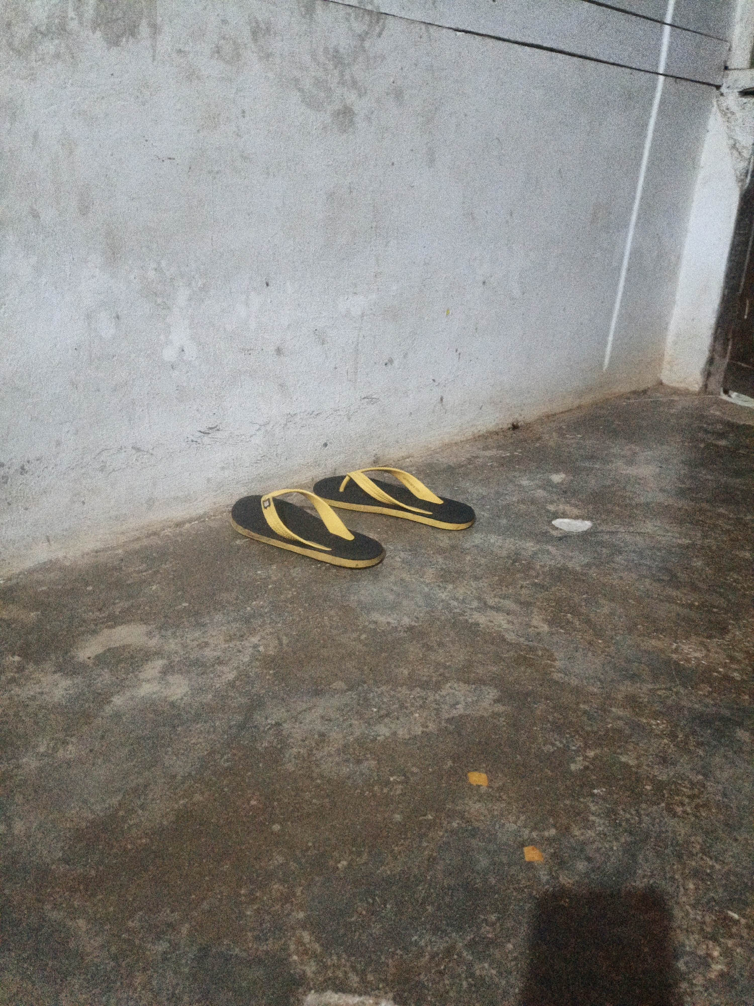 A pair of black and yellow flip-flops are placed on a dirty, wet concrete floor in front of a rough, gray concrete wall. The scene appears to be outdoors, possibly in a low-income area or alleyway within Meunasahuket, Indonesia. There are a few small pieces of debris scattered on the floor, including a white crumpled piece of paper and two small yellow squares. The lighting is dim, and the overall impression is one of neglect.