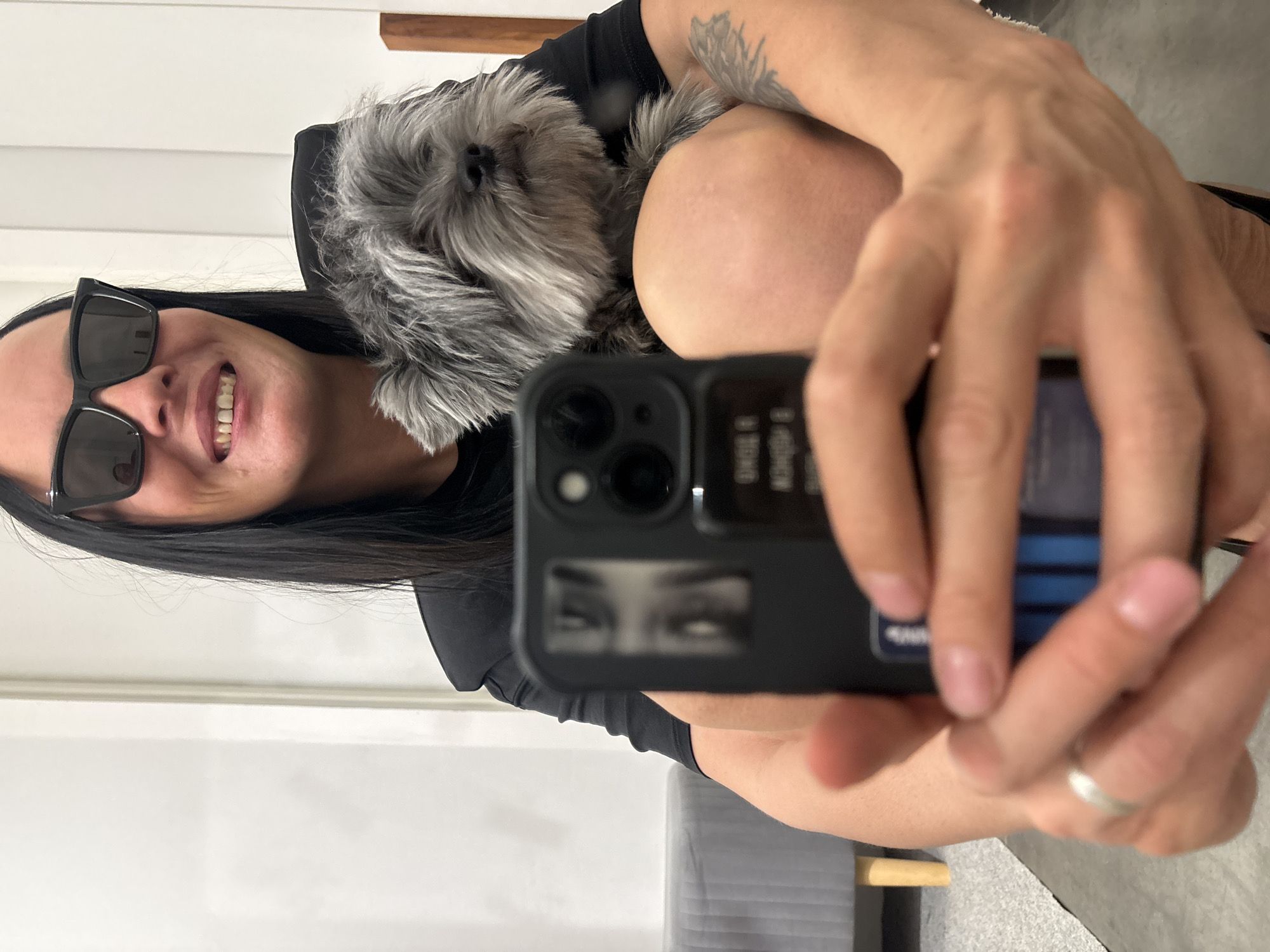 A woman with black sunglasses smiles into a smartphone camera, holding the phone with her right hand while a gray, long-haired dog is nestled against her left shoulder. She is wearing a black shirt. The background includes a white wall and a gray chair. The location is unknown and there is no evidence it is in Chaikovsky, Russia.