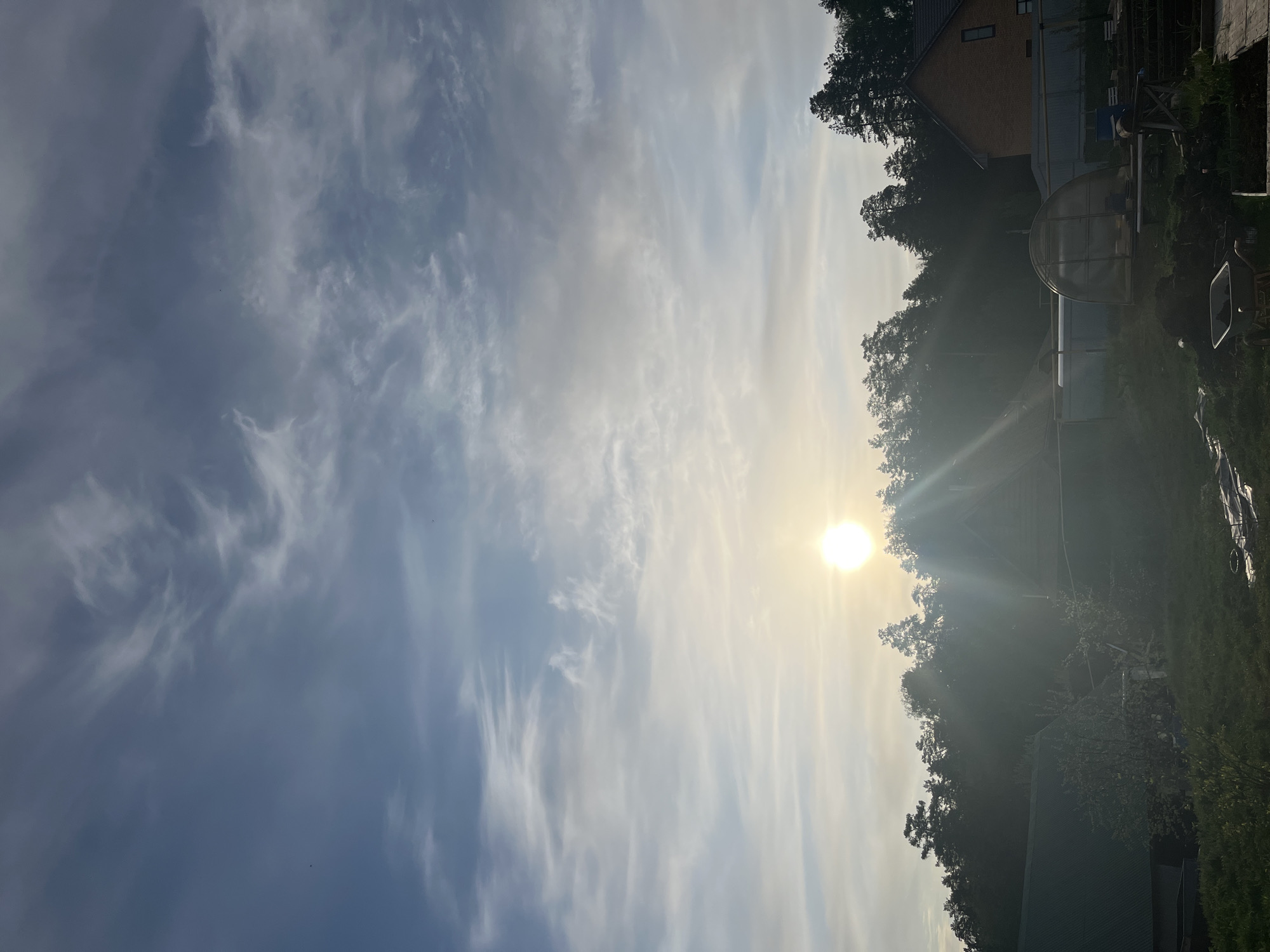 The image shows a view in Chaikovsky, Russia, under a bright, sunny sky with light, streaky clouds. The sun is positioned near the tree line, causing lens flare. Silhouetted trees and structures, including a greenhouse, are visible in the lower right portion of the image. The overall composition suggests a rural or suburban setting. A brick building is visible among the trees. The image appears to be taken facing southeast towards the location of the sun.