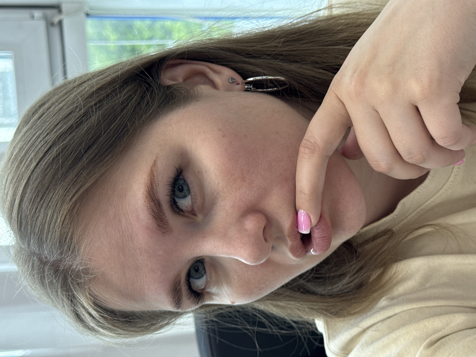 A young woman is facing the camera. She has fair skin, light brown hair, and blue eyes. Her finger is touching her lips. She wears hoop earrings and a small heart-shaped earring in her ear. The environment is indistinct, but a window is visible in the background, suggesting an indoor setting. The location, Perm, Russia, is not discernible from the image.