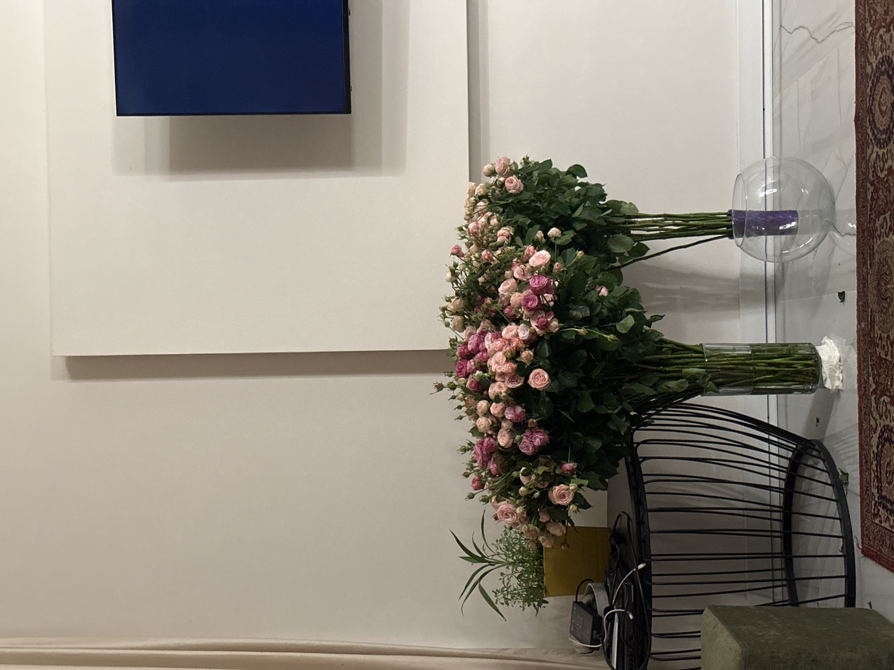 The image displays an indoor scene, primarily featuring a large arrangement of flowers against a light-colored wall. The original image is oriented sideways, but I will describe it as if it were upright.
On the left side of the frame, a dark blue rectangular object, possibly a screen or a piece of minimalist art, is mounted on a plain light beige wall. It casts a soft shadow below it.
Dominating the center and right side of the image is a substantial collection of fresh flowers, specifically spray roses, featuring various shades of pink, from light blush to deeper magenta, interspersed with abundant green foliage. These flowers are divided into two main bunches, each held in a clear glass vase. The top vase is a spherical or bulbous glass container, holding one bunch of roses whose stems are wrapped with a purple material. Below it, a tall, cylindrical clear glass vase holds another large bunch of roses, with white crumpled material visible at the bottom of the vase, likely to support the stems or absorb water.
To the bottom right, a piece of black, open-slat or wire furniture, possibly an accent chair or basket, is partially visible. Next to this, closer to the foreground, a small green plant with slender leaves is situated in a yellow container. Nearby, various electronic cables and a black power adapter are visible, suggesting an area for charging devices or powering electronics. In the very bottom right corner, a portion of a light green or olive-colored upholstered stool or ottoman is present.
In the far background, on the right side of the image, a vertical white architectural element, possibly a door frame or wall corner, is seen. Further to the right, a section of a patterned rug, possibly an oriental or Persian design with dominant red, brown, and cream tones, is visible, adding a touch of texture and color to the otherwise neutral palette.
The lighting appears to be indoor and diffused, creating soft shadows. There is no visible text in the image. The overall impression is one of an indoor setting, possibly a living room or a display area, with fresh flowers arranged decoratively.