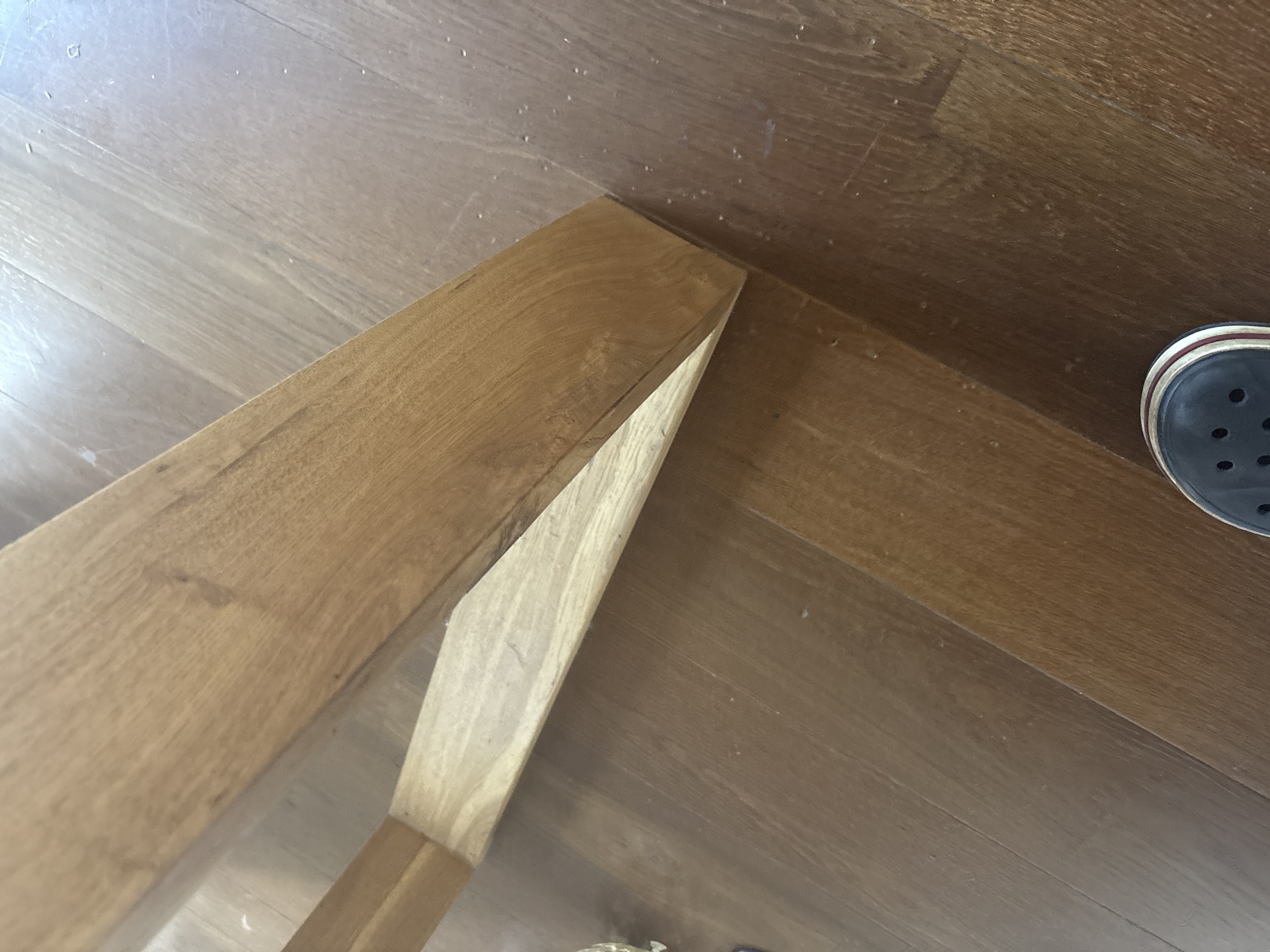The image shows a close-up view of a wooden floor and a wooden furniture leg. The floor is made of wooden planks with a visible wood grain pattern. The furniture leg is made of light-colored wood and has a trapezoidal shape. A black shoe with white and red accents is visible in the lower right corner of the image. The overall scene appears to be indoors, possibly in a residential setting. There is no indication of the location being in the city of Ореховка, Russia.