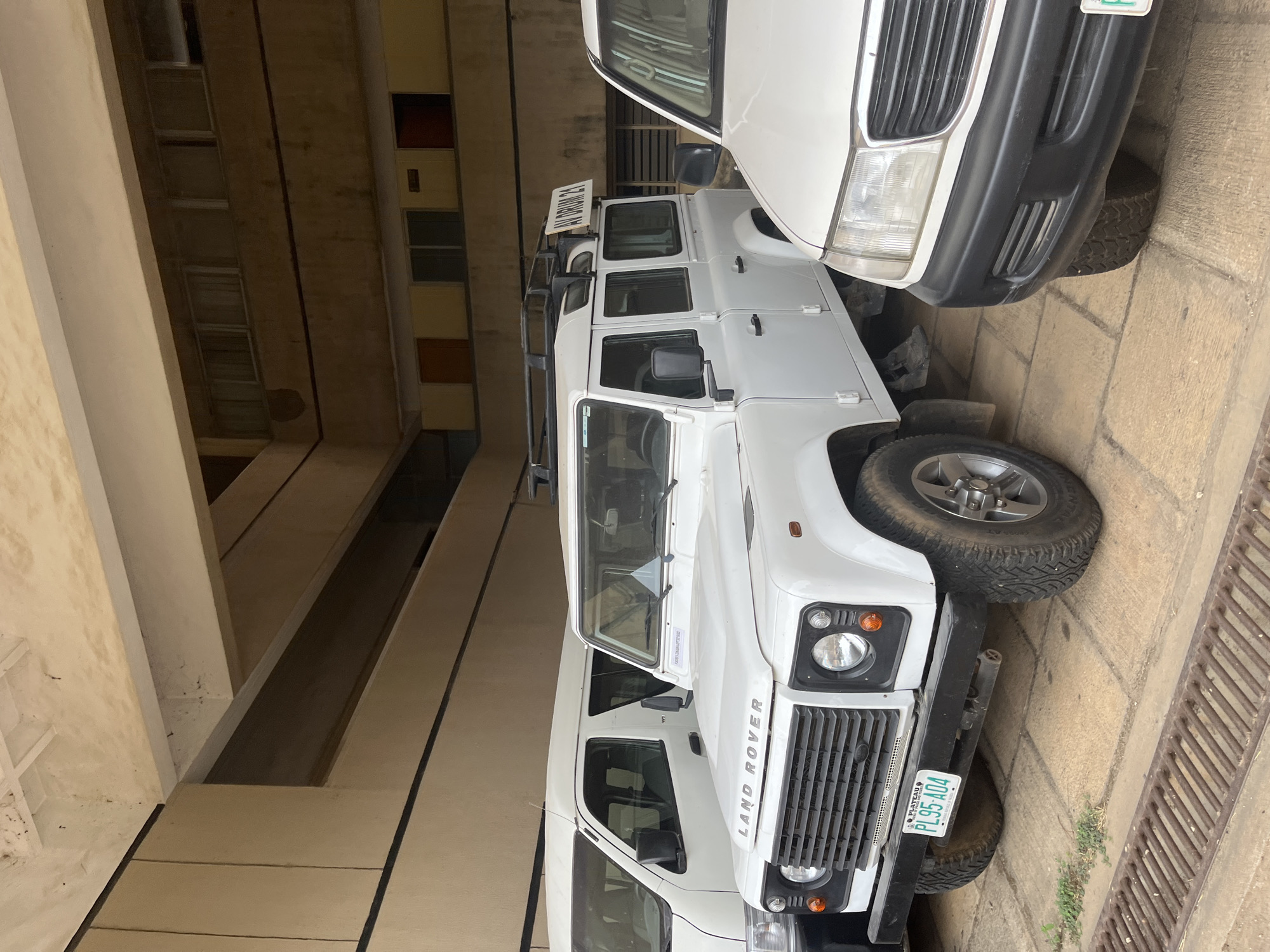 A white Land Rover Defender, bearing license plate "PL95-A04", is parked in a covered area in Jos, Nigeria. It is positioned next to another white vehicle, partially visible in the upper right of the frame. The Defender is oriented with its front towards the viewer. The surrounding environment features a building with light-colored structural supports and visible windows, indicating an urban or semi-urban setting. The ground is paved and includes a drainage system. There are no people visible in the image.