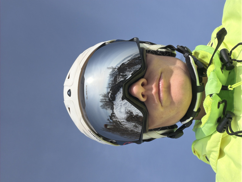 A close-up portrait depicts an individual wearing ski equipment against a clear blue sky. The person wears a white Atomic-branded ski helmet and silver-mirrored Blizzard ski goggles, which reflect a panoramic view of a snow-covered mountain landscape. The reflection includes bare trees, exposed rocky terrain, a bright sun glare, and a distant ski lift structure. The lower section of the individual's face, encompassing the nose, mouth, and chin, is visible beneath the goggles. They are dressed in a neon yellow-green ski jacket featuring black drawstrings and toggles around the collar. The scene depicts a high-altitude outdoor environment consistent with a ski resort in the city of Zelenaya Polyana, Russia.