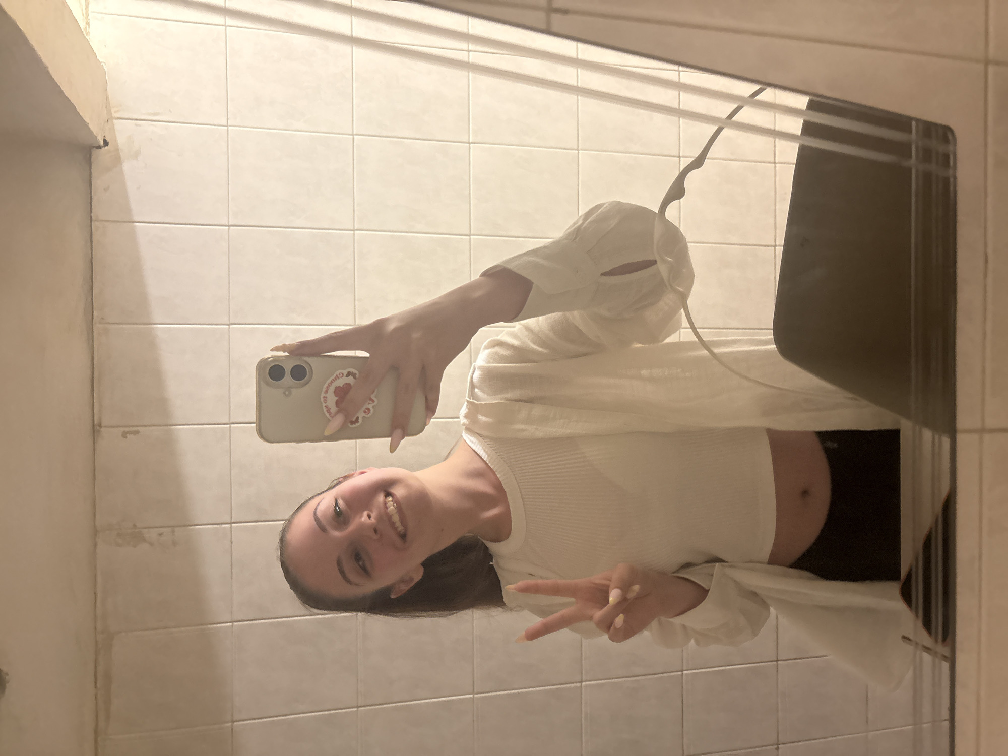 A young woman with dark hair pulled into a ponytail takes a selfie in a bathroom. She is wearing a white ribbed crop top and a white open cardigan. She is smiling and holding up two fingers in a peace sign with her left hand. Her right hand is holding a light-colored phone, which has a red sticker on the back. The bathroom has tiled walls and a mirror that reflects the scene. The location is not specified in the image or context.