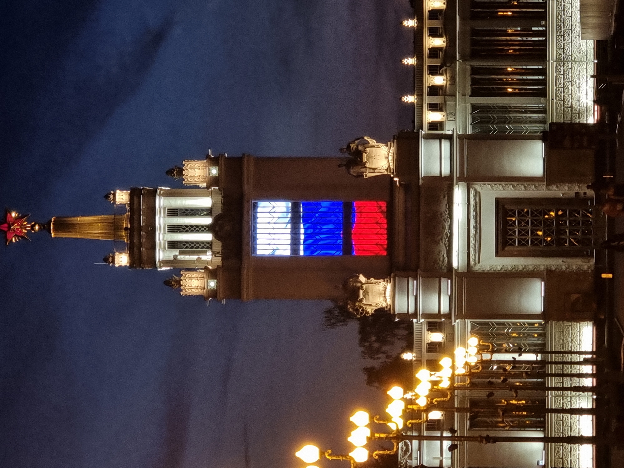 The image shows the Moscow Main Trade Show pavilion at night, illuminated by artificial lights. The pavilion's tower is lit up, displaying the Russian flag colors in a large window. The red star atop the tower is also lit. In the foreground, a row of ornate streetlights shines brightly, illuminating the surrounding area. The sky is dark, indicating it is nighttime, with some cloud cover visible. The overall scene depicts a monument illuminated at night.