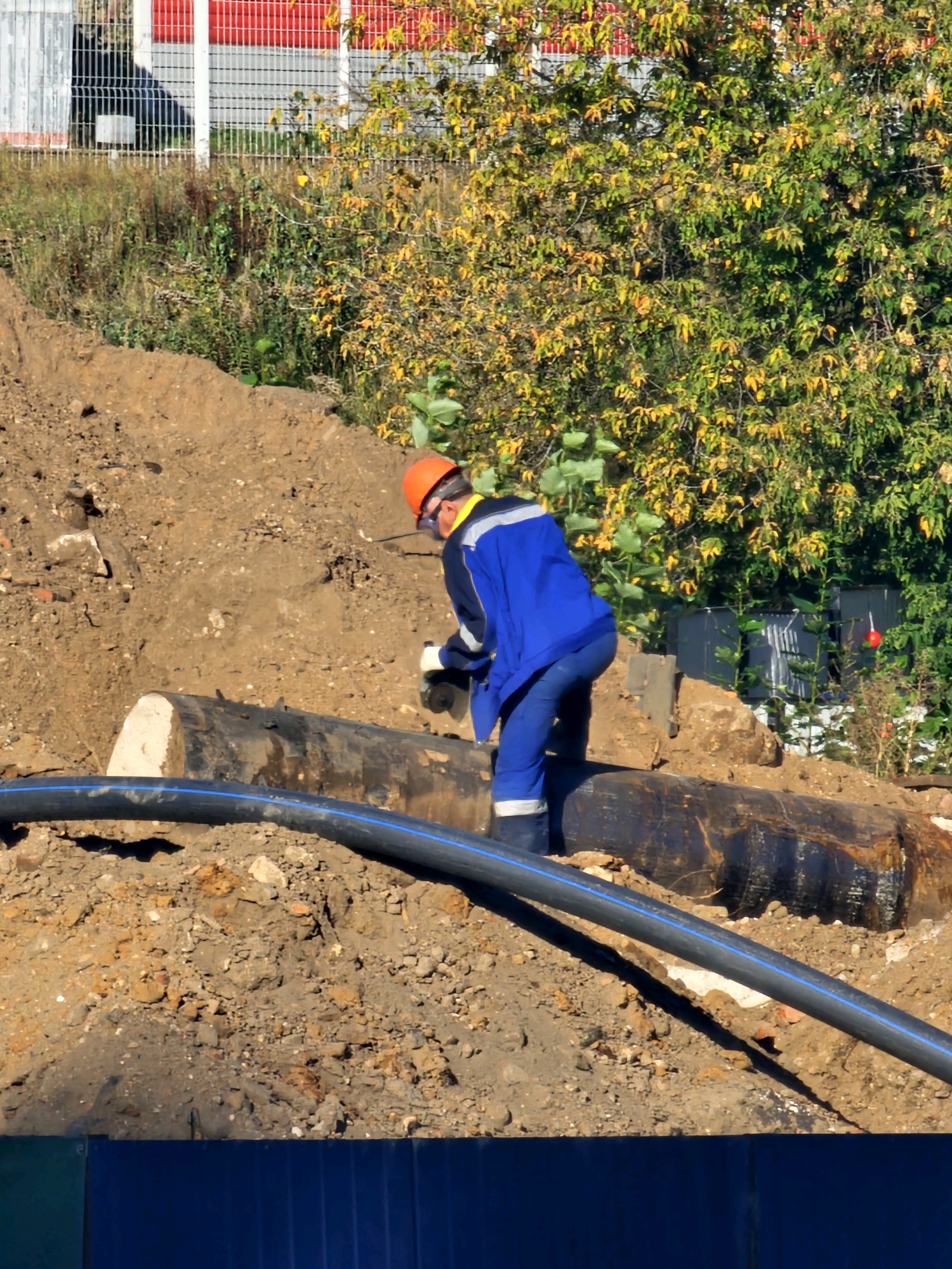 A construction worker in a blue uniform and orange hard hat is operating a grinder on a large, dark-colored pipe laid on a dirt embankment. A black plastic pipe with a blue stripe runs across the foreground. Behind the worker, a dense tree with yellow and green leaves stands against a backdrop of a building with red and white vertical stripes and a fence. The scene appears to be a construction site, possibly in Moscow, Russia, characterized by exposed earth and building materials.