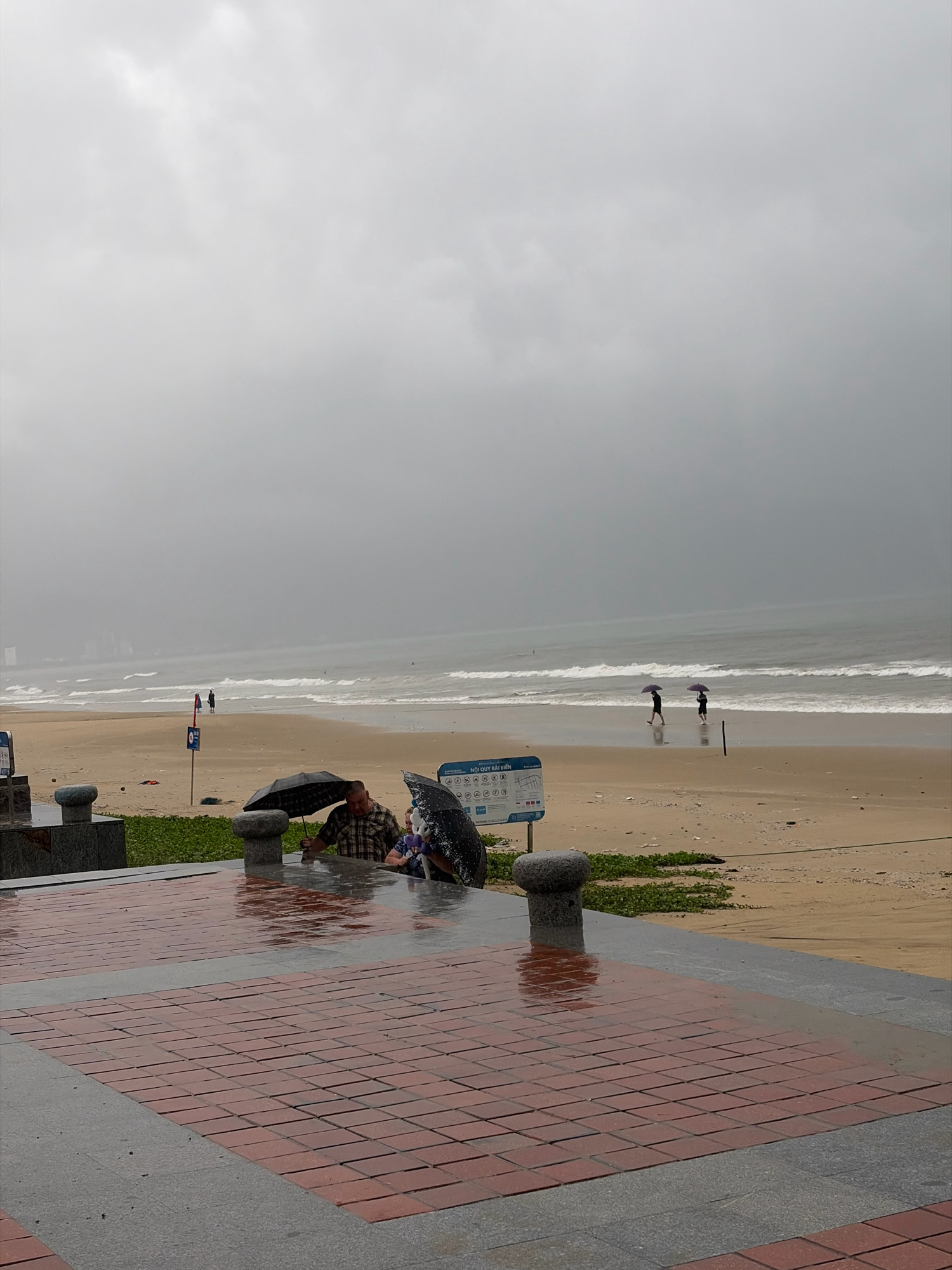The image depicts a gray, overcast day on a beach in Đà Nẵng, Vietnam. The foreground shows a wet, red brick and gray stone promenade, suggesting recent or ongoing rain. Two people are visible under umbrellas, one man in a plaid shirt and another person partially obscured, both sitting near a dark stone bollard. To the left, another bollard is visible. Behind them, the sandy beach is mostly empty, with some green vegetation and scattered debris. Two signs are posted on the beach; one, a blue and white informational sign, is to the right of center. Further out, the ocean is visible with moderate waves, and two figures are walking on the wet sand near the water's edge, each holding a dark umbrella. In the distance, faint outlines of buildings suggest an urban setting beyond the beach. The overall atmosphere is somber and damp, indicative of bad weather.
