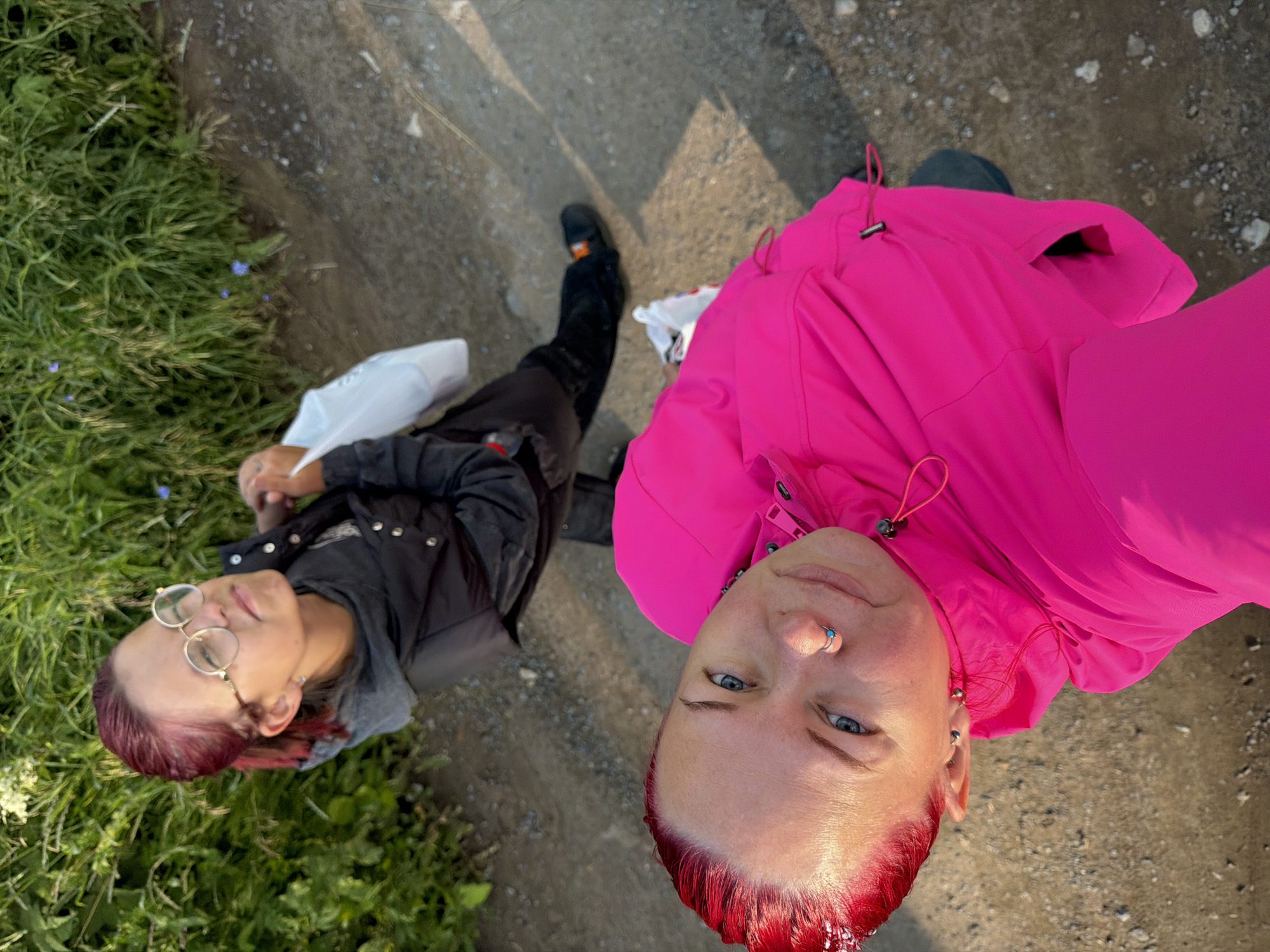 An overhead shot shows two individuals outdoors on a dirt path bordered by lush green grass. The person in the foreground, closer to the camera, has bright red hair and wears a vibrant pink jacket. They are looking up at the camera with a slight smile and have a nose ring. Their head is tilted upwards, and the shot is taken from their perspective.
In the background, further up the path, a second person with deep red hair is visible. They are wearing a black puffer vest over a grey hooded jacket and round glasses. This individual is also looking upwards, with a more neutral expression, and is holding a white plastic bag.
The environment suggests a natural setting, possibly on the outskirts of Chaikovsky, Russia, where the path leads alongside a vegetated area. The lighting indicates it's daytime, with shadows cast on the path, suggesting the sun is at an angle.