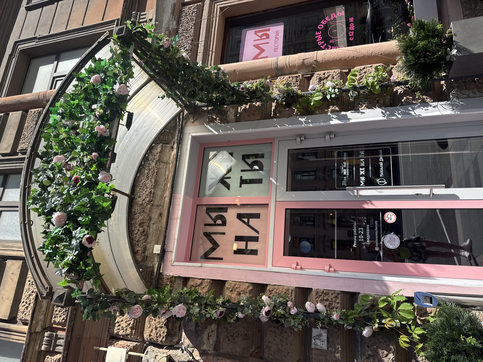 The image shows the exterior of a restaurant or cafe in Saint Petersburg, Russia, decorated with artificial ivy and pink roses. The building has a stone facade and an arched entrance with a clear canopy. The entrance features a pink frame with large windows displaying text in black Cyrillic letters. On the left window, the words "МЫ НА" are visible. On the right window, the word "ТЫ" is visible. The door to the left has text that reads "Режим работы: 10-23 ежедневно" (Opening hours: 10-23 daily) and "restoranmyzhenaty". The door to the right has pink text reading "МЫ РЕСТОРАН" and "быстрые обеды с 12 до 16" (quick lunches from 12 to 16). A person, reflected in the glass door, is wearing a plaid skirt and white sneakers. The overall impression is of a charming and inviting establishment.