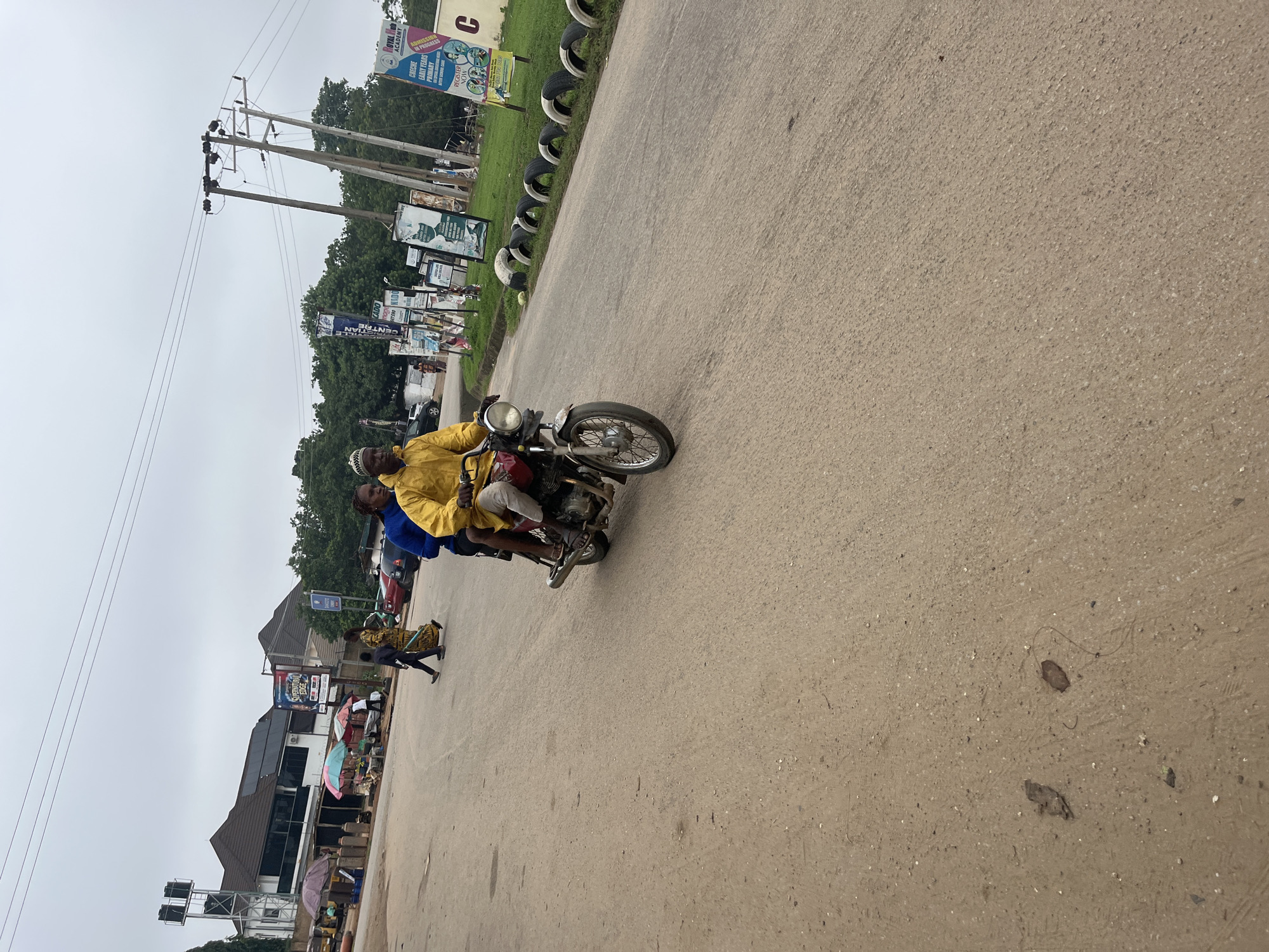 Two individuals are riding on a red motorcycle on a paved road. The person in the front is wearing a yellow hooded jacket and a patterned cap, while the person behind is wearing a blue top. The motorcycle has a visible headlight and is moving forward. The road appears to be in a commercial or residential area, with buildings and street vendors visible in the background. Several utility poles with overhead wires are present along the right side of the road, and some of the poles are adorned with posters and advertisements, including one for "Royal Hub Academy." The environment suggests an outdoor setting in a city with moderate traffic and pedestrian activity. The ground is a mixture of asphalt and dirt, with some debris scattered about. The sky is overcast, indicating a cloudy day. The scene depicts a typical street view in Abuja, Nigeria.